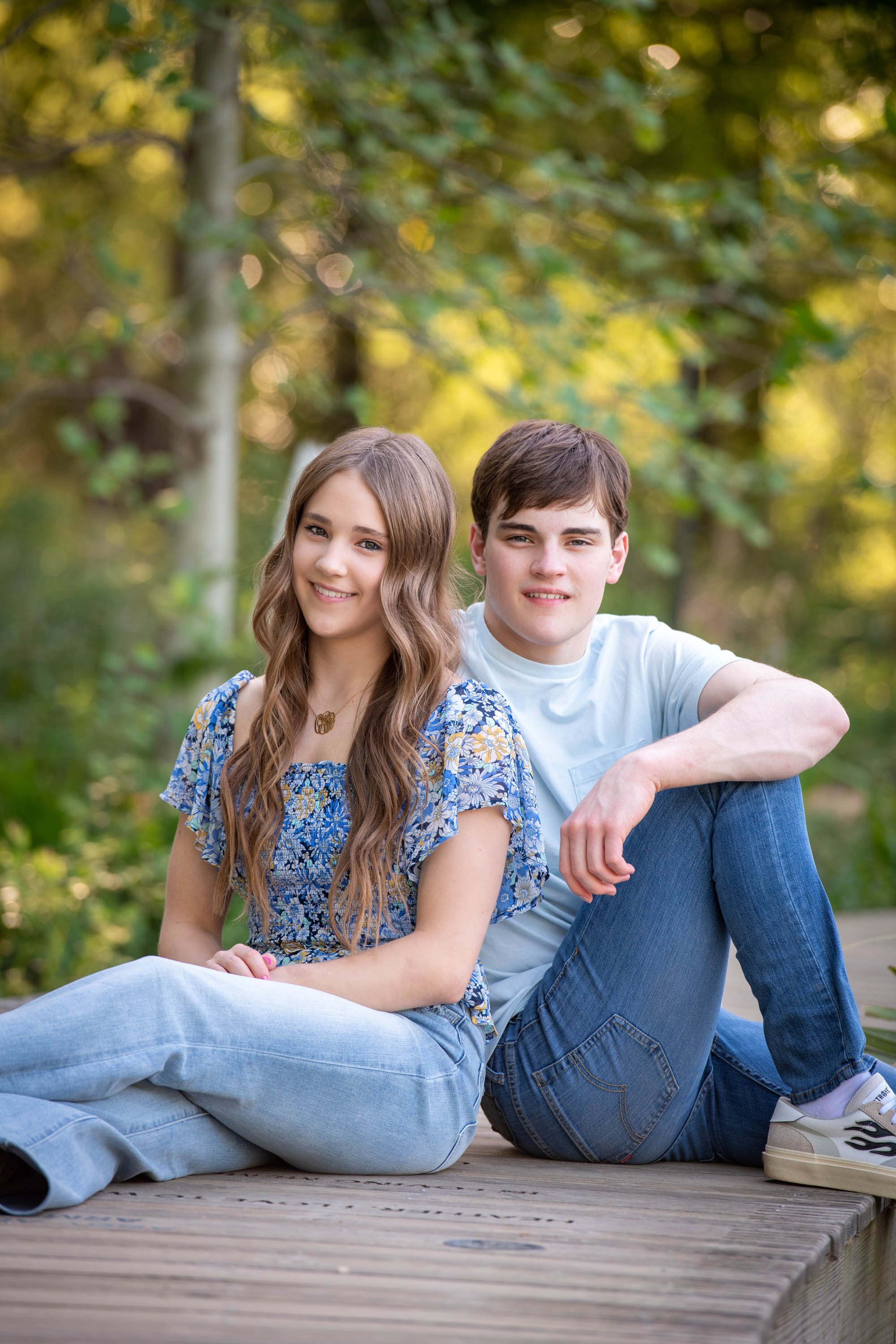 A young woman with long, wavy hair and a young man with short hair sitting together on a wooden deck outdoors, surrounded by green trees, smiling at the camera.