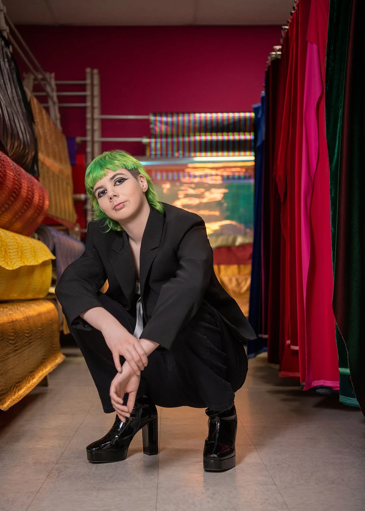 Person with bright green hair, wearing a black suit and black platform boots, crouching in a colorful fabric store aisle.