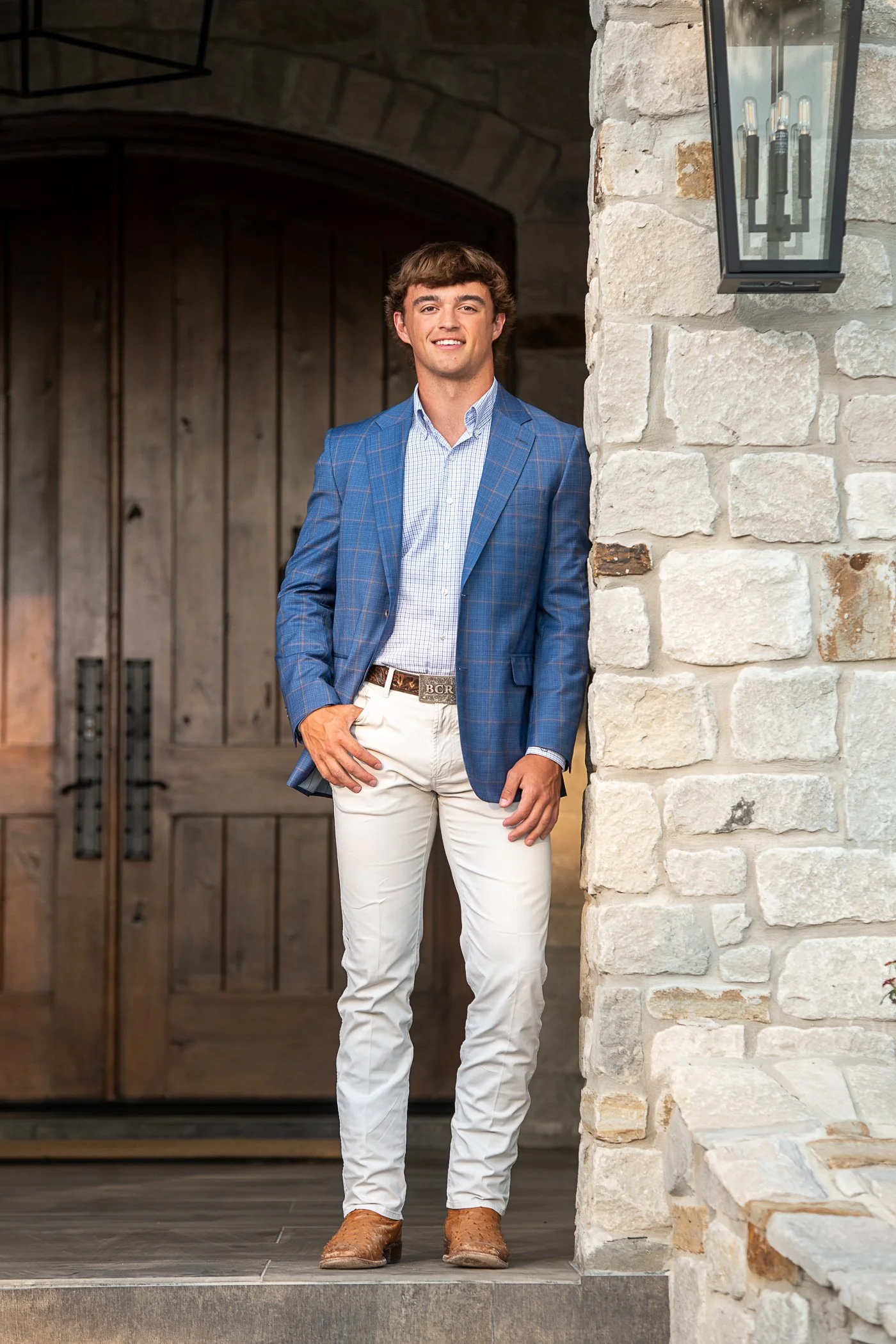 A young man in a blue plaid blazer, light blue shirt, and white pants stands on a front porch with a stone wall and dark wooden archway behind him. He is smiling and has one hand on his hip, wearing brown boots.