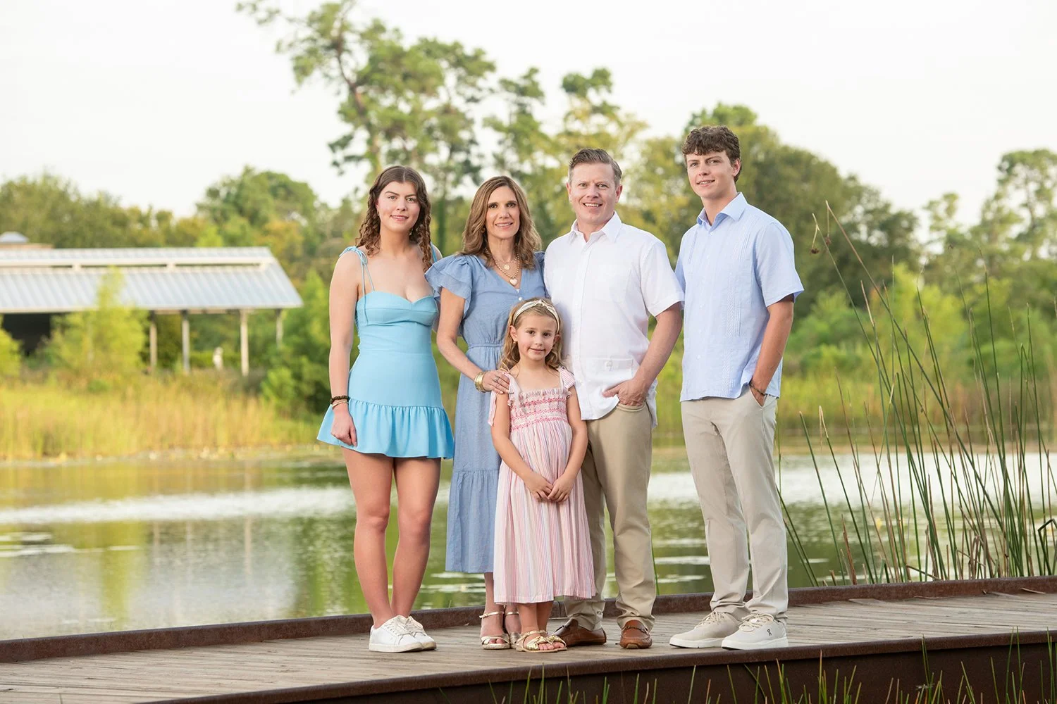 A family standing on a wooden platform beside a lake in a park, with trees and a building with a metal roof in the background. The family includes a young women, one woman, one young girl, one man, and one teenage boy, all dressed in summer clothing.