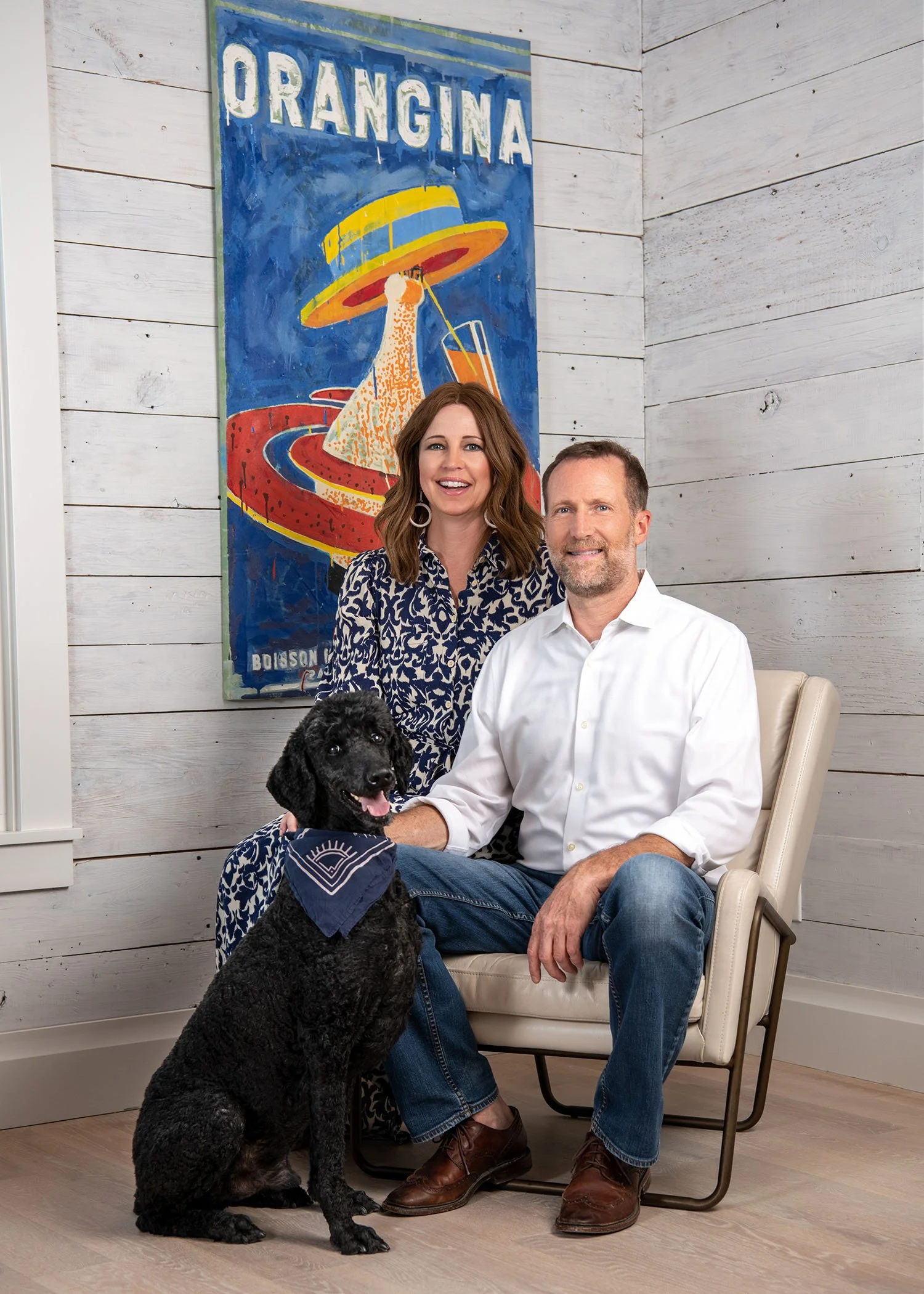 A man and woman sitting together in a room with white wood-paneled walls, smiling at the camera. The woman has shoulder-length brown hair and is wearing a patterned dress, while the man has short hair and a beard, wearing a white shirt and jeans. A b