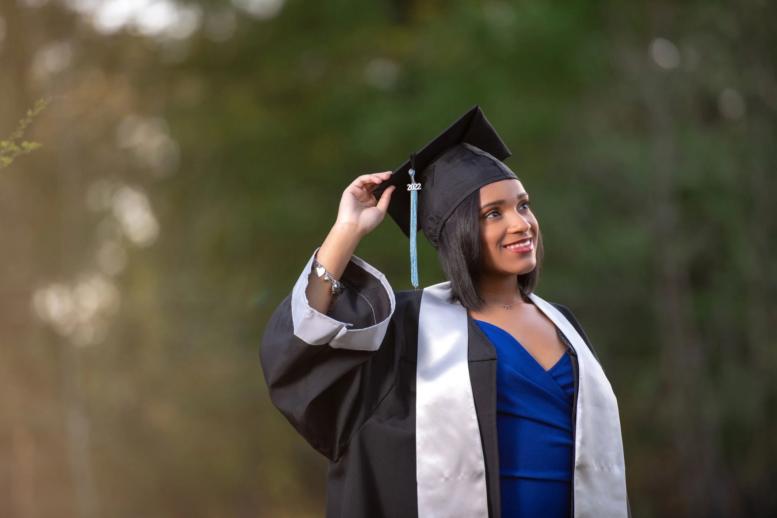 A young woman in a graduation cap and gown smiling outdoors, holding her cap with the year 2022 on the tassel.