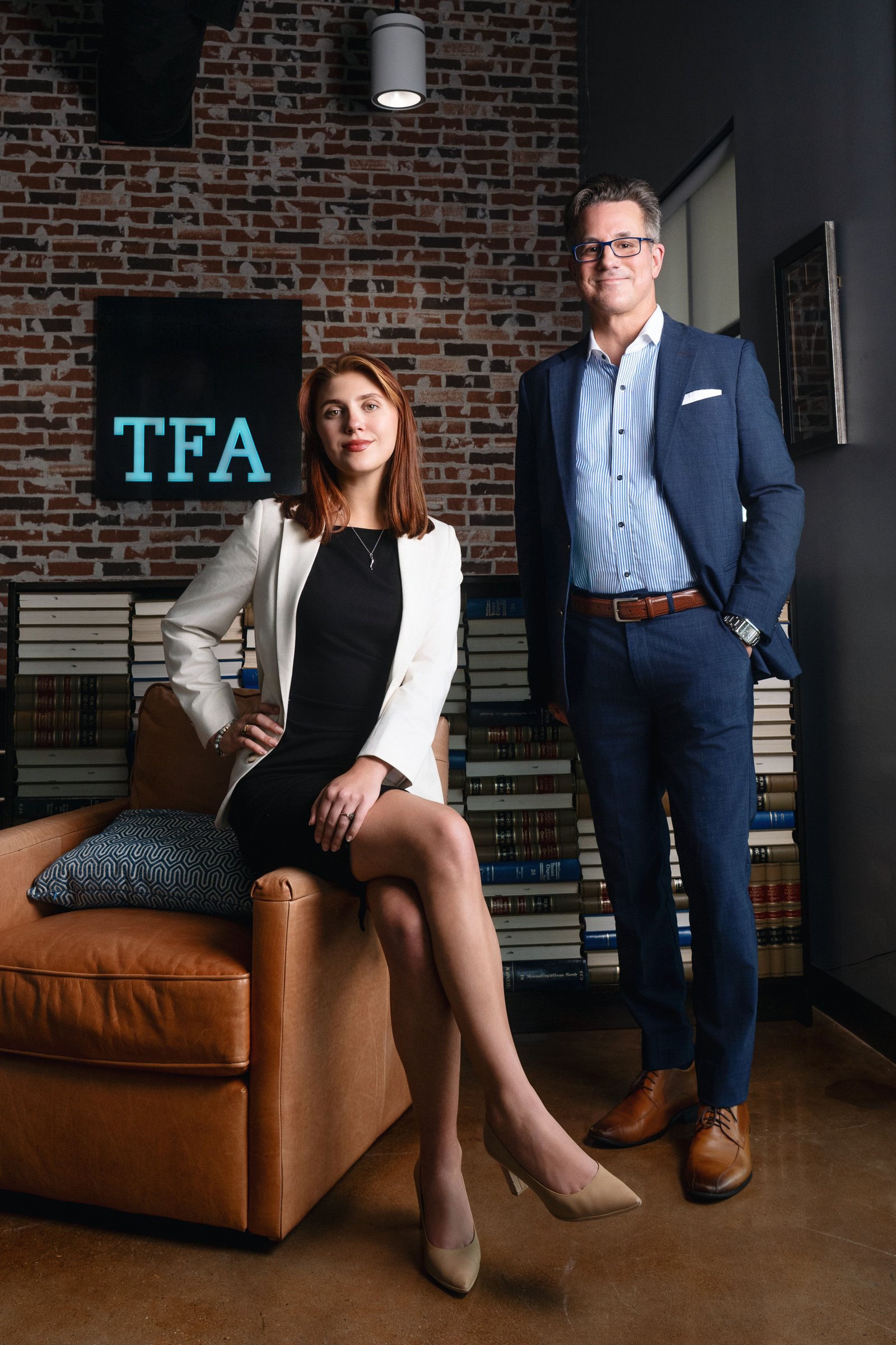 A woman in a white blazer and black dress sitting on a tan leather couch with a pillow, and a man in a blue suit standing beside her in a room with a brick wall, bookshelf, and a sign that reads 'TFA' in illuminated letters.