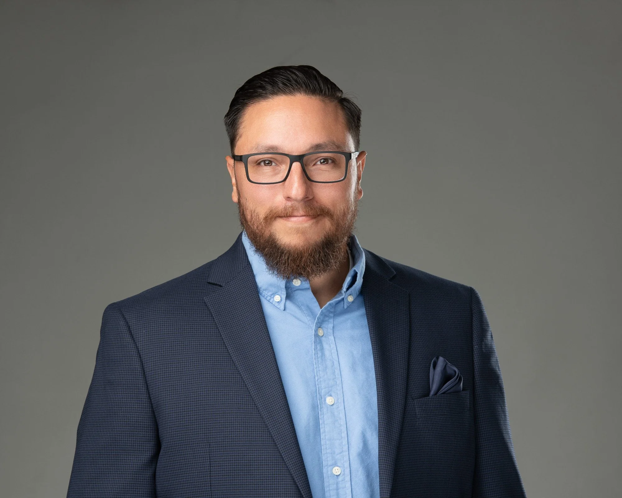 Man with dark hair, glasses, a beard, wearing a navy suit jacket, light blue dress shirt, and a pocket square, posing against a gray backdrop.