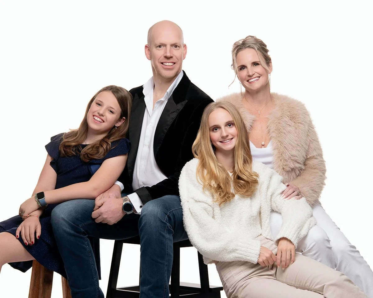 Family of four sitting together on chairs against a white background, smiling for the camera. The group includes a man, a woman, and two young girls.
