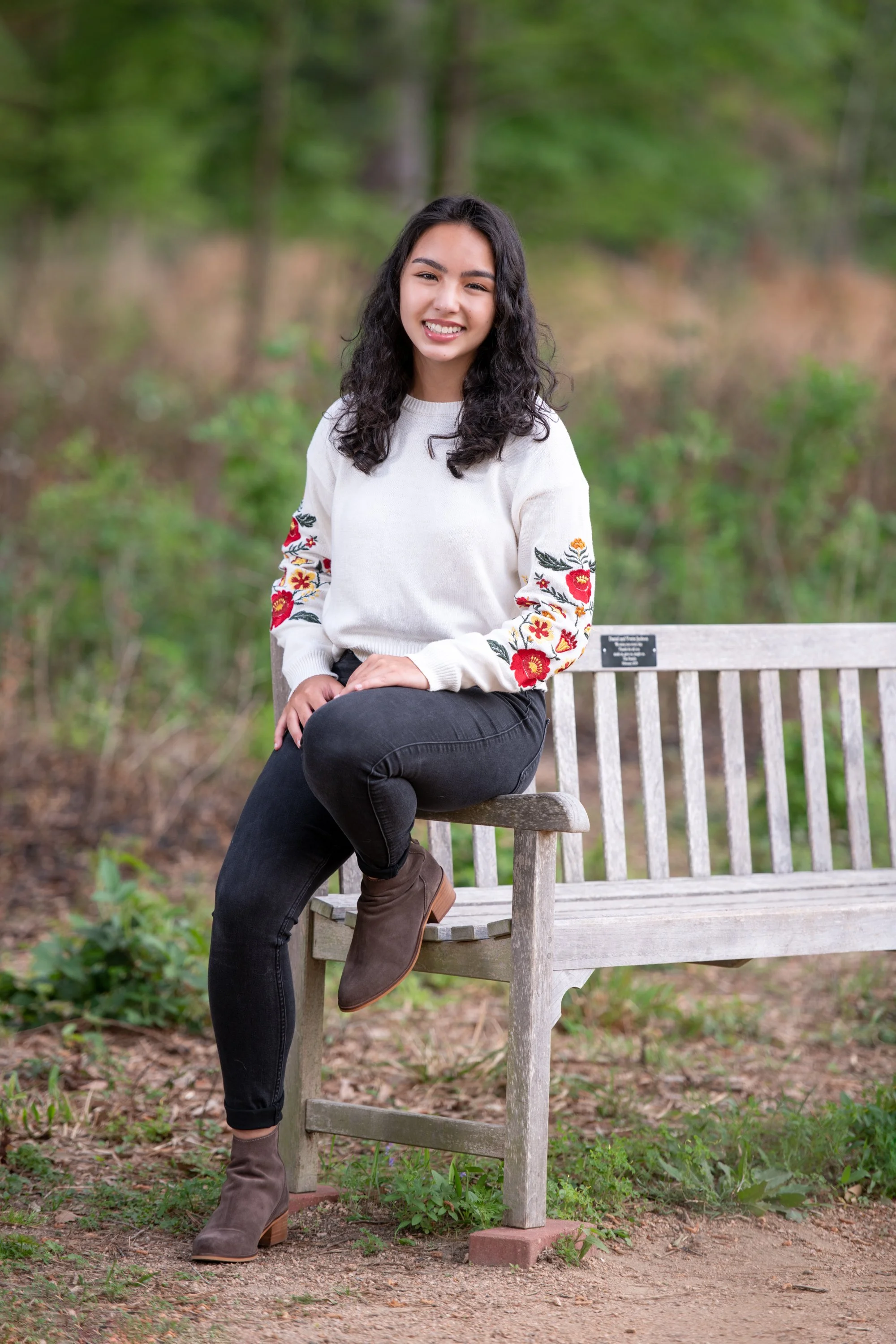 A young woman with dark, curly hair, sitting on a wooden park bench outdoors in a green, wooded area, smiling at the camera.