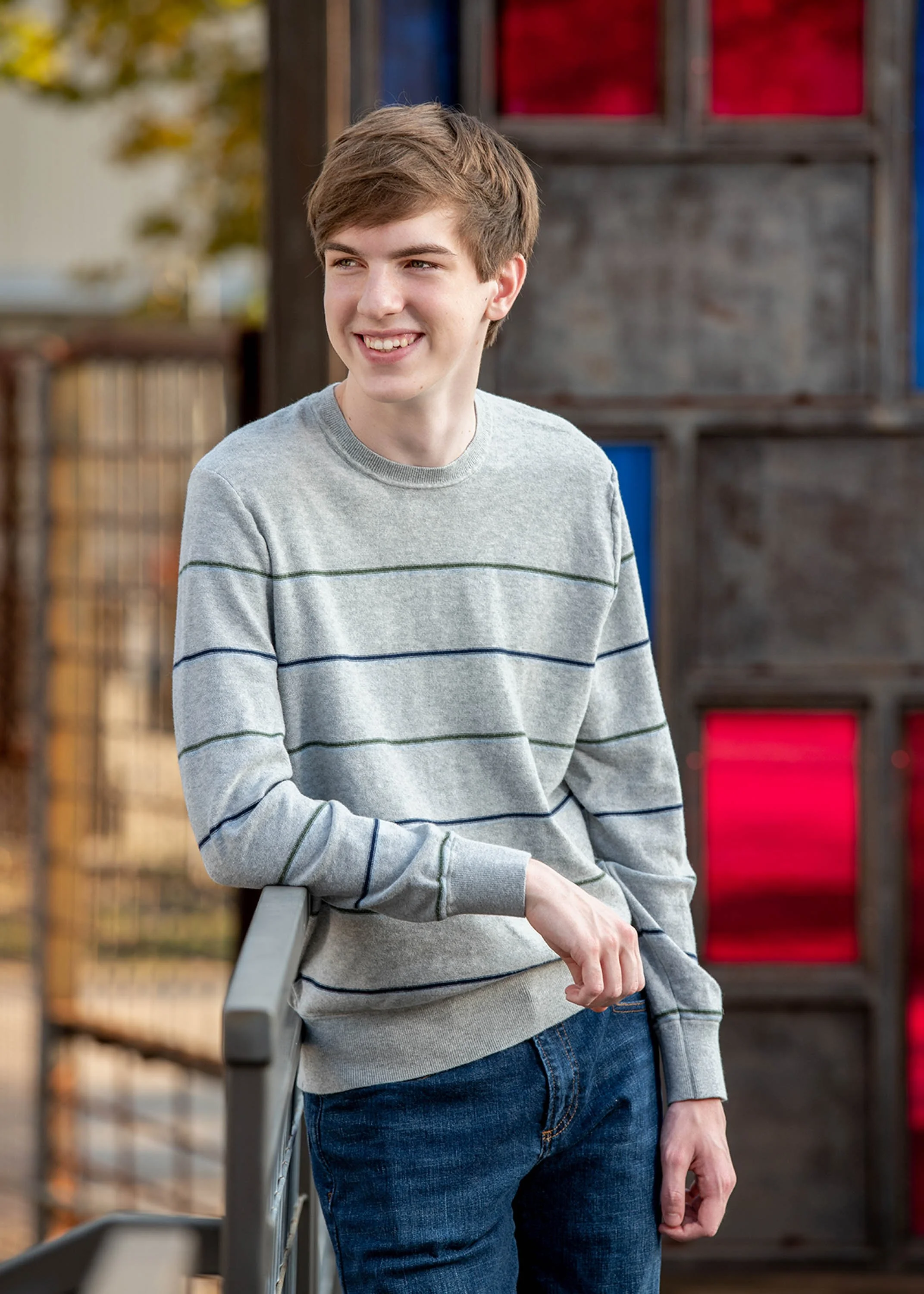 A young man with brown hair wearing a gray sweater with black and green horizontal stripes standing outdoors, leaning on a gray railing with his left arm, and smiling. In the background, there are autumn trees and a wooden structure with red and blue