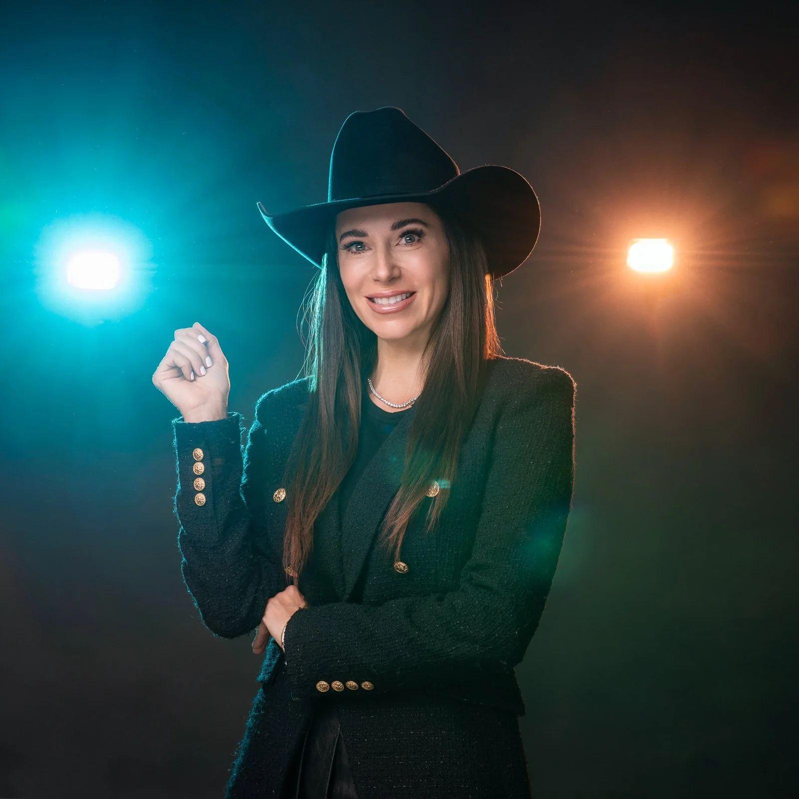 A woman wearing a black cowboy hat and a black jacket with gold buttons, smiling, with colorful lights in the background.