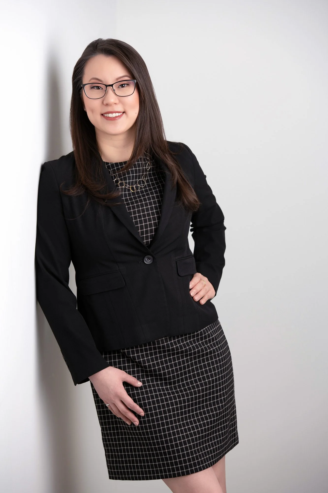 A professional woman with glasses, wearing a black blazer and a checkered skirt, standing against a plain white background.
