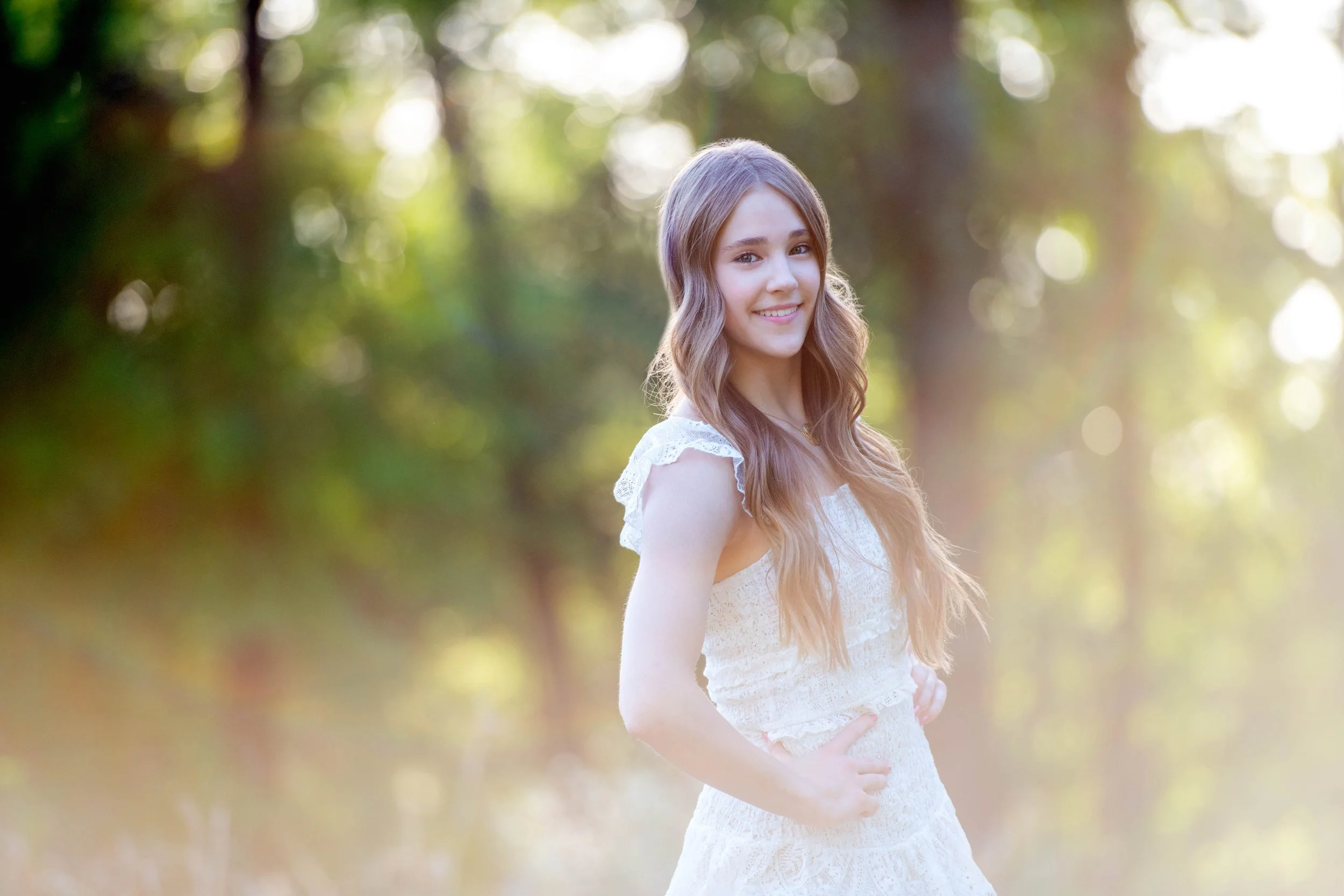 A young woman with long wavy brown hair, wearing a white lace dress, standing outdoors in a forested area with sunlight filtering through the trees, smiling gently at the camera.