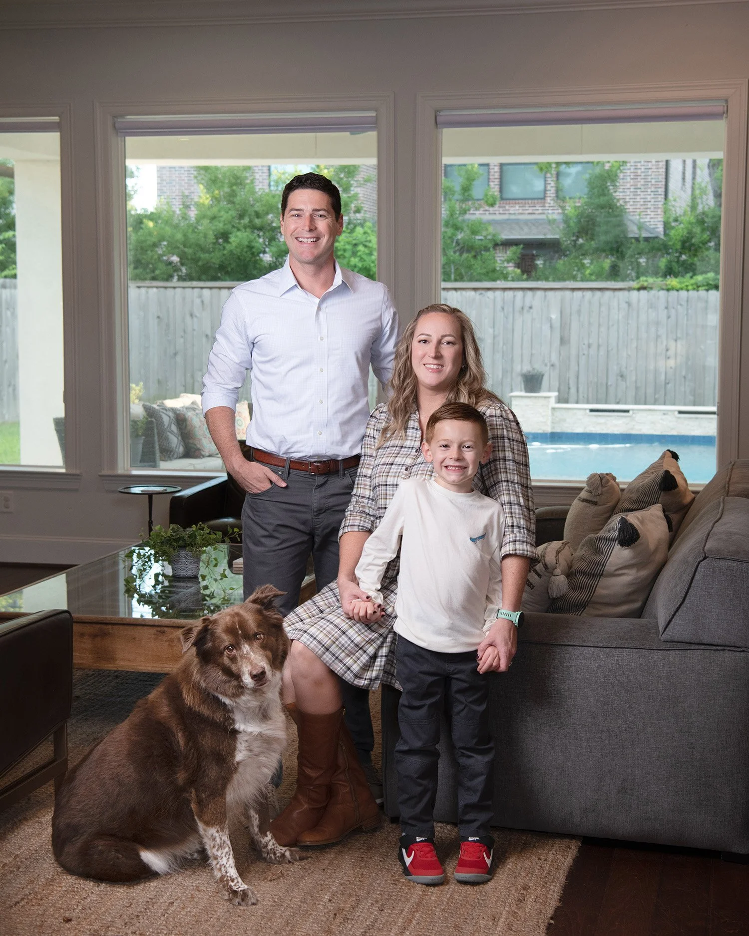 Family of four, two adults and two children, along with a brown dog, posing inside a living room with large windows showing a backyard with a swimming pool and trees.