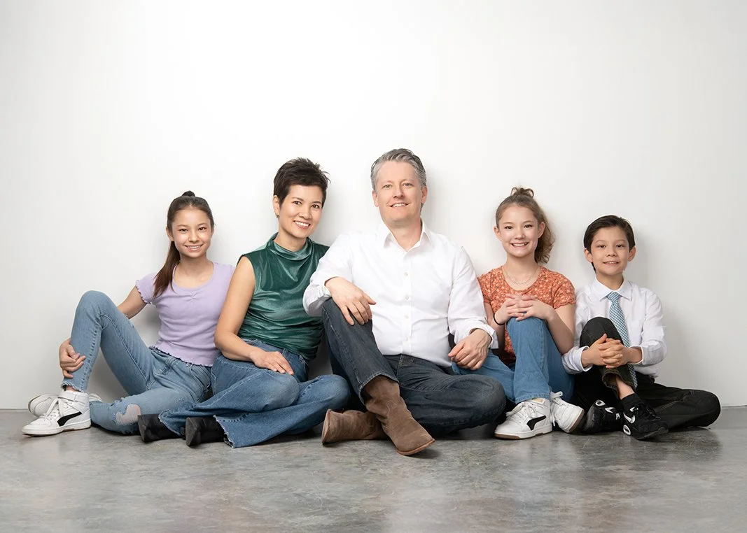 A family of five, including a man, a woman, and three children, sitting on the floor against a white wall, smiling at the camera.
