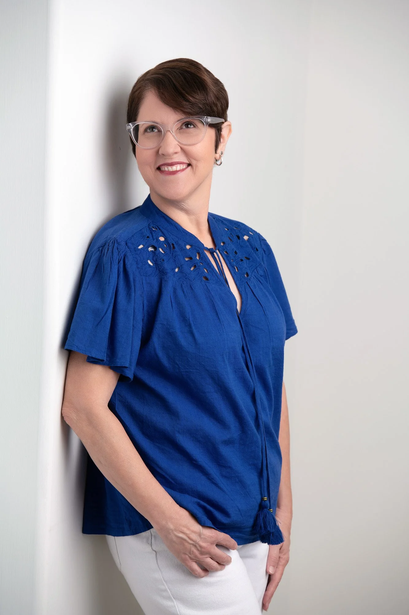 A woman with short brown hair and glasses leaning against a white wall, wearing a bright blue blouse with lace and a tie detail near the neckline, and white pants, smiling and looking off to the side.