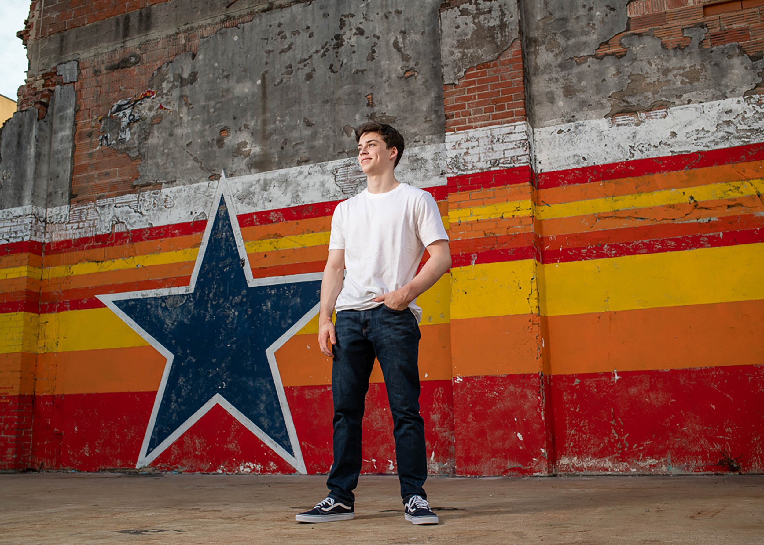 A young man in a white t-shirt and jeans standing in front of a colorful, weathered wall with a large star painted on it, featuring red, orange, yellow, and white stripes.