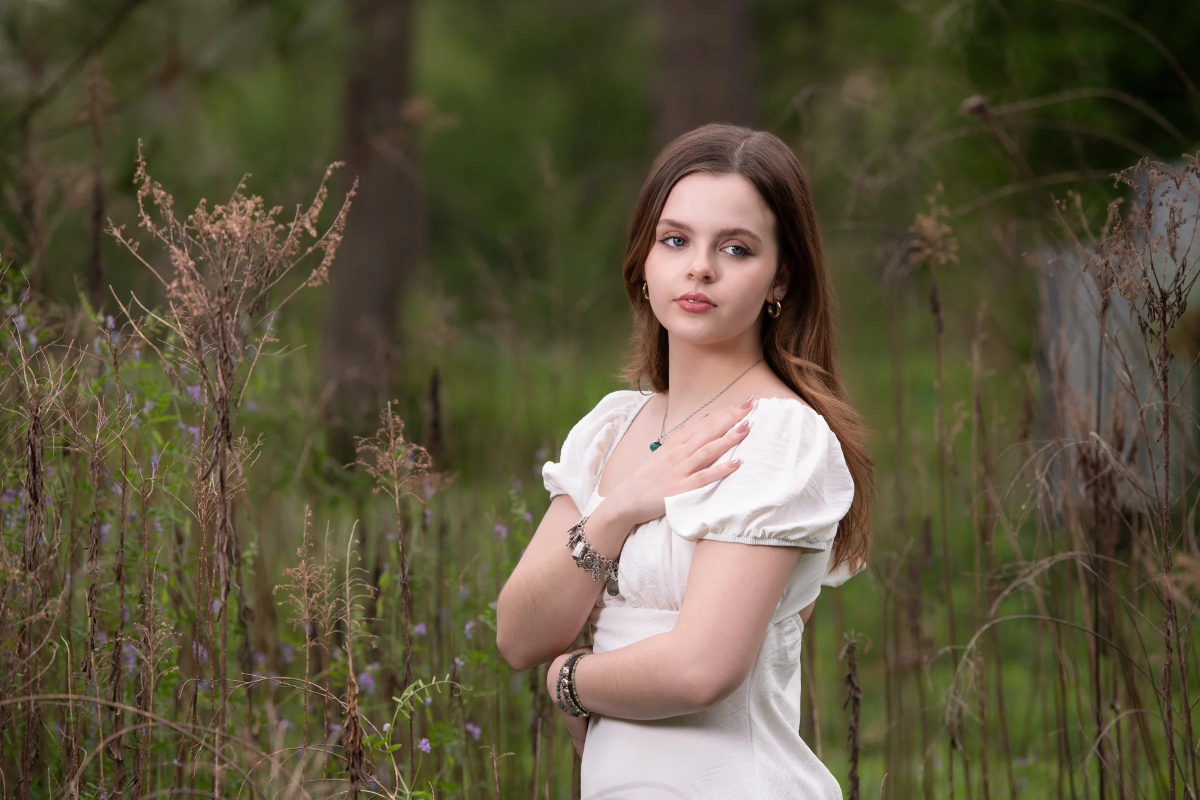 A young woman with long brown hair, wearing a white dress and jewelry, posing outdoors in a natural setting with green trees and brown plants.