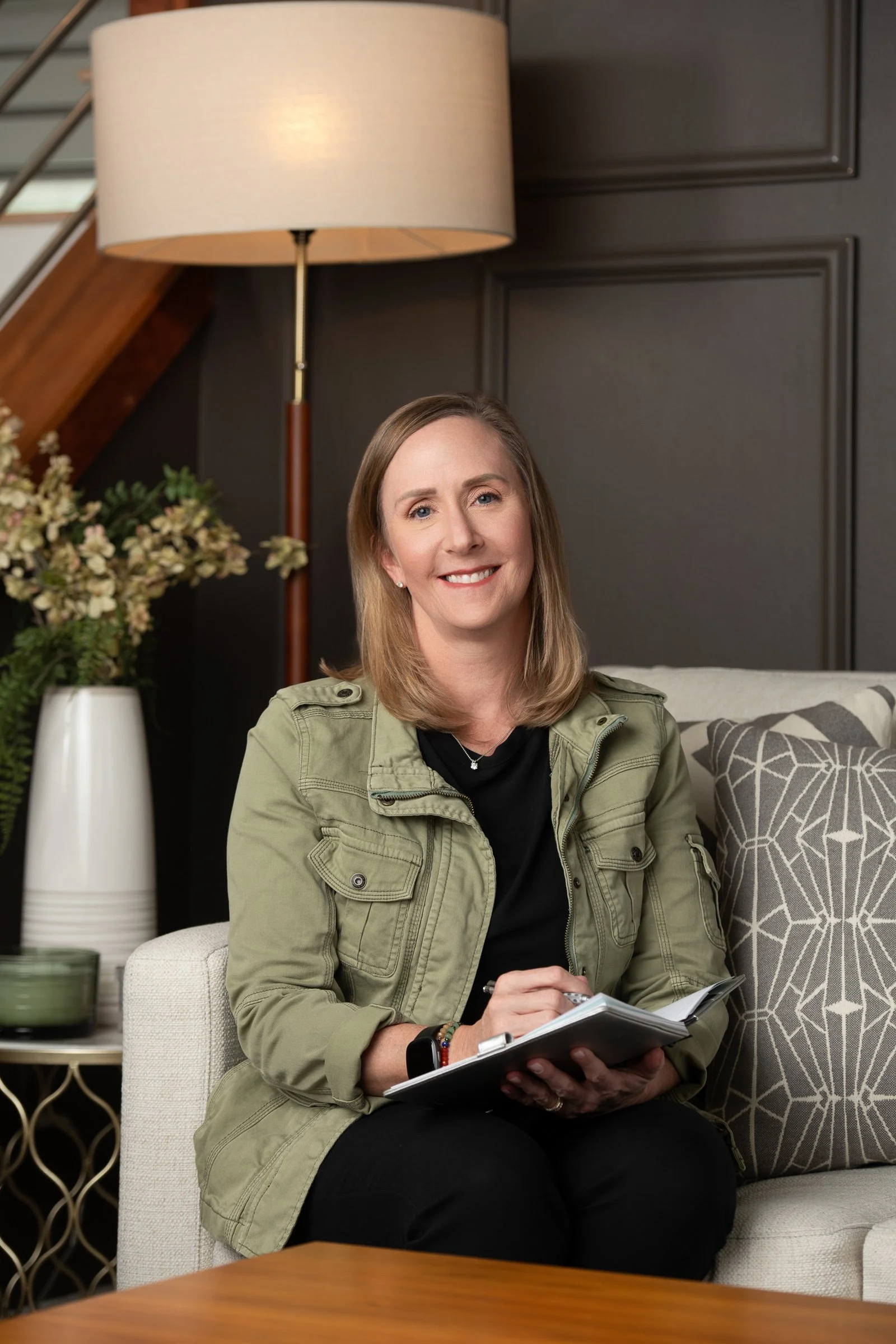 A woman with shoulder-length blonde hair, wearing a light green jacket and black shirt, sits on a beige armchair in a living room. She is smiling and holding a notebook and pen. Behind her is a dark wall with wood paneling, a large beige lampshade, a