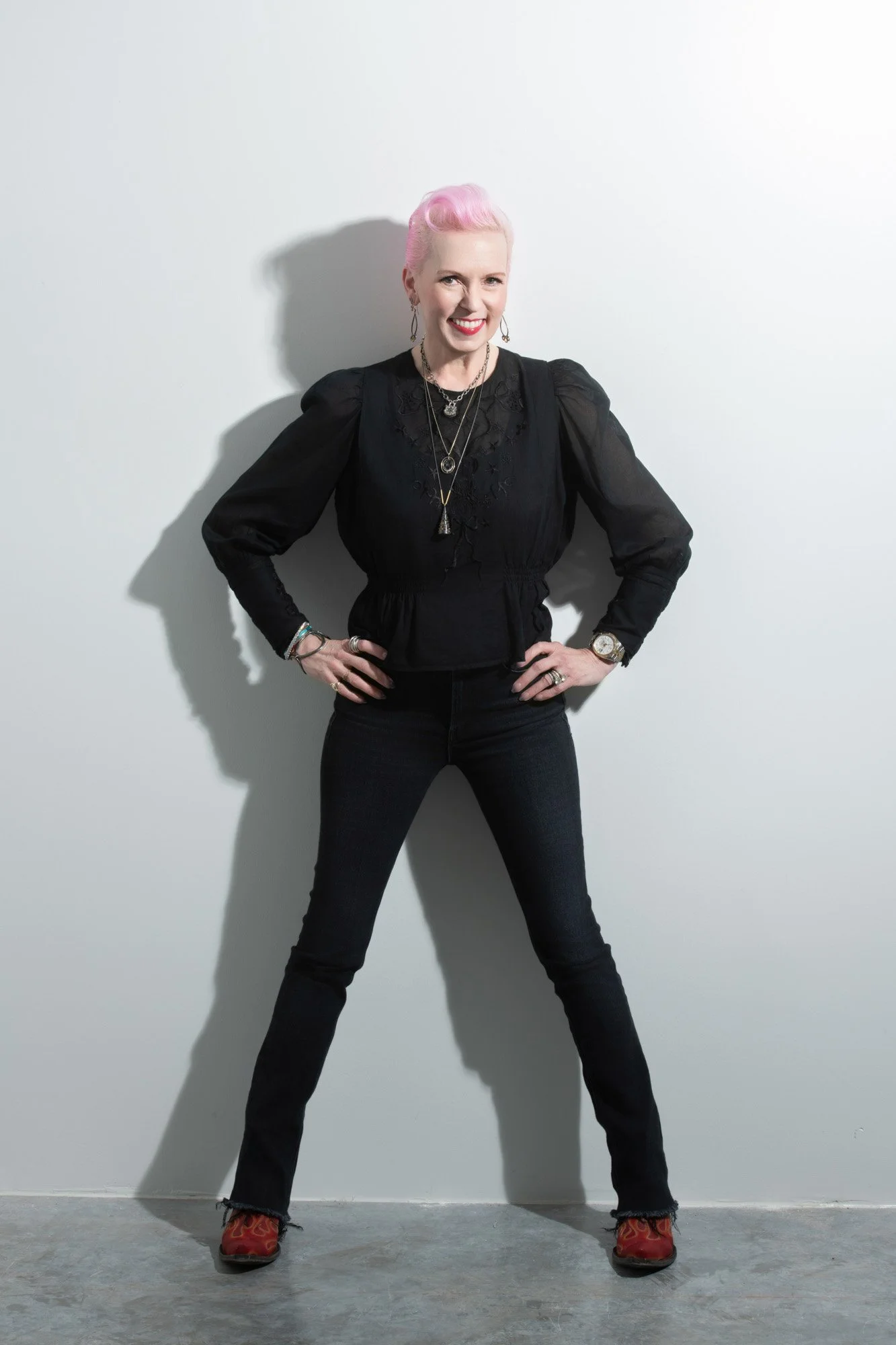 A woman with short pink hair, wearing a black blouse with puffed sleeves and black jeans, stands against a plain white wall. She's smiling with her hands on her hips, wearing jewelry, a watch, and red cowboy boots with flames.