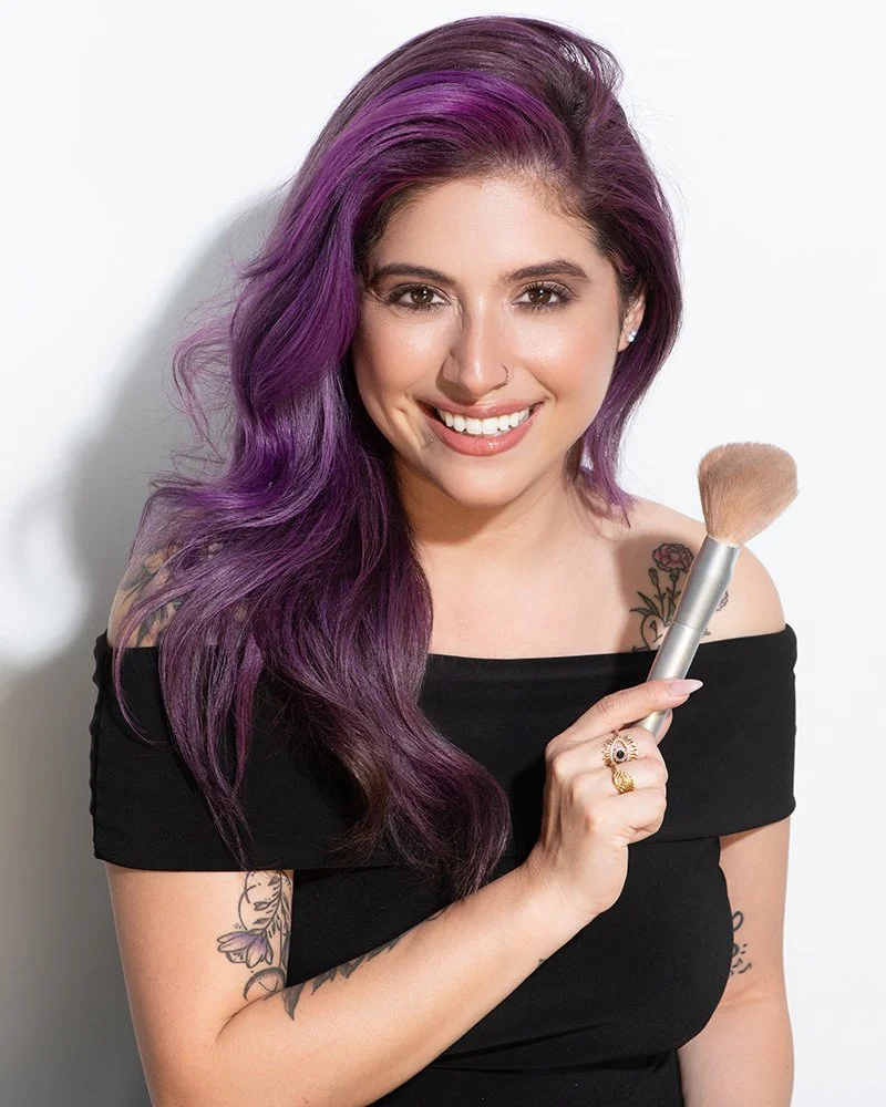 A woman with purple hair holding a makeup brush, smiling, wearing a black off-shoulder top, and showing tattoos on her arm and shoulder.