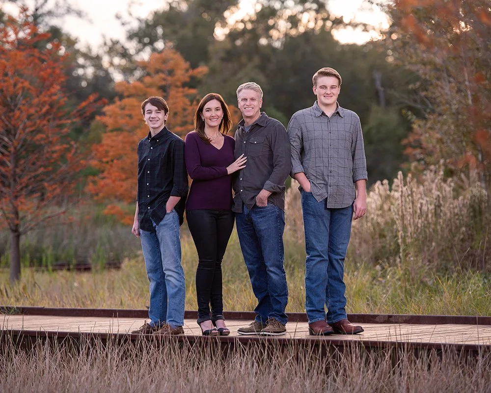 Family of four standing on a wooden bridge outdoors with fall foliage in the background.