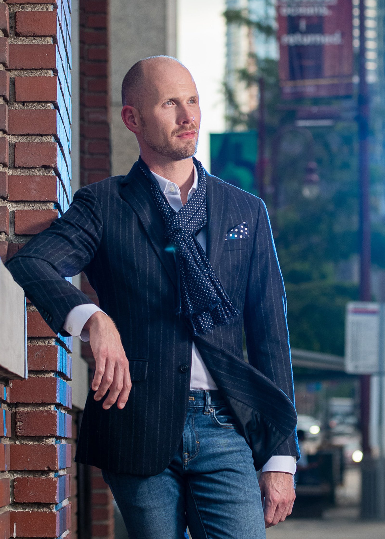 A man with a shaved head and beard leaning against a brick wall outdoors, wearing a dark pinstripe suit jacket, a white shirt, and a blue patterned scarf, with a cityscape and blurred background.