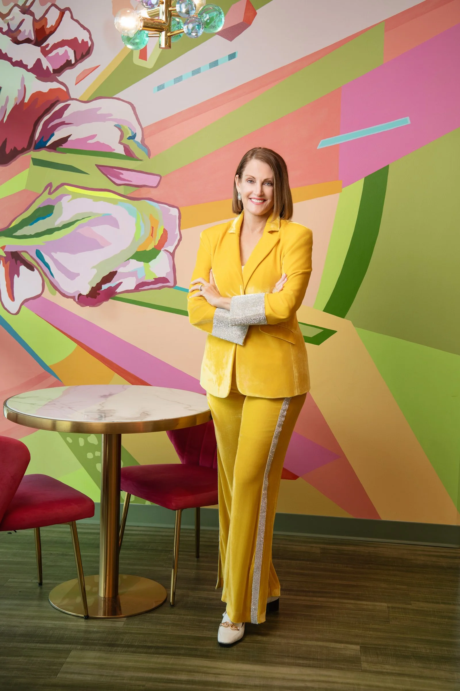 A woman in a yellow suit with silver accents on the sleeves and a stripe down the side of her pants, standing in front of a colorful, stylized floral mural, with her arms crossed and smiling.