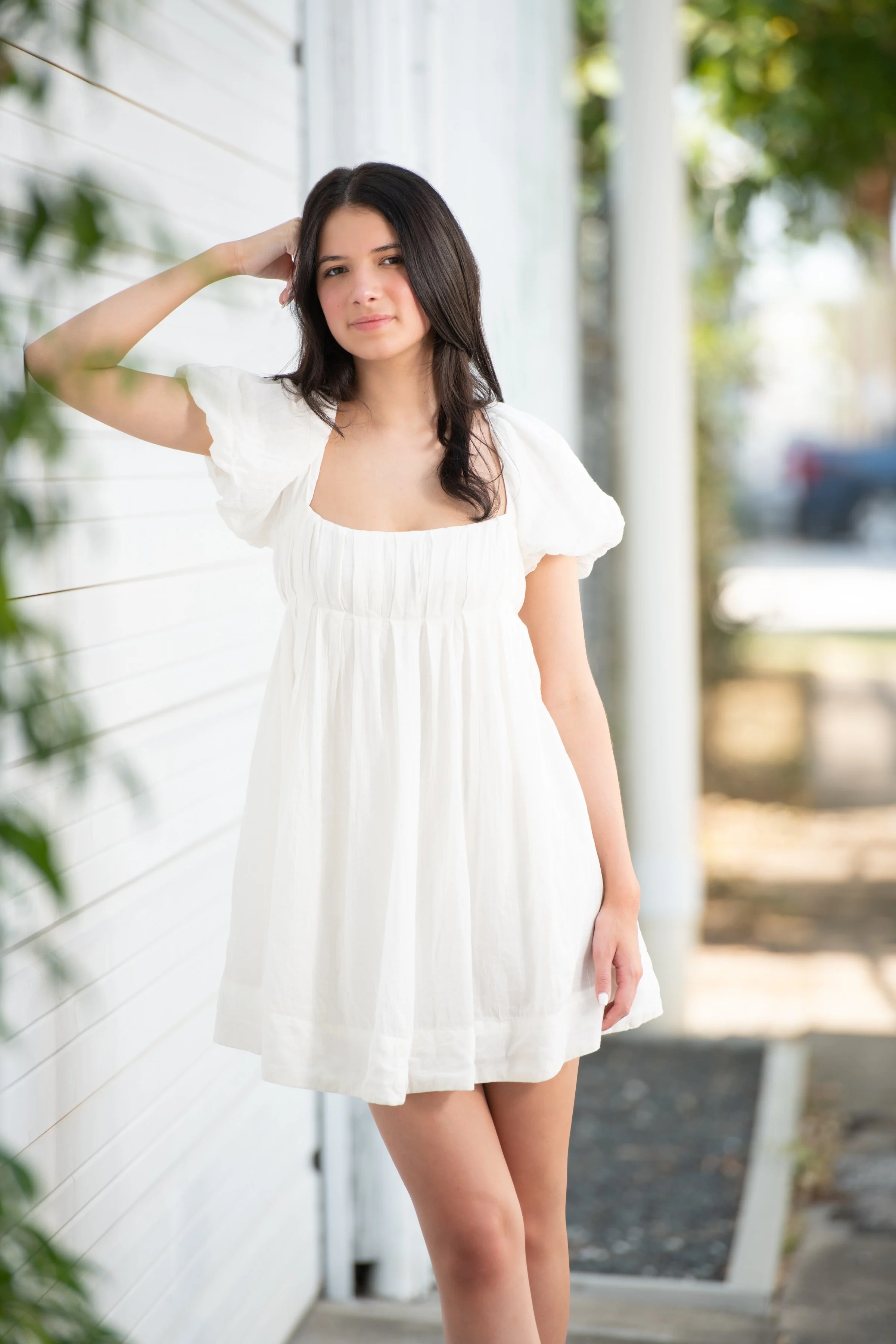 A young woman with dark hair wearing a white dress standing outside near a white wall and a glass door, leaning against the wall with one arm raised and hand on her head, with blurred trees and cars in the background.