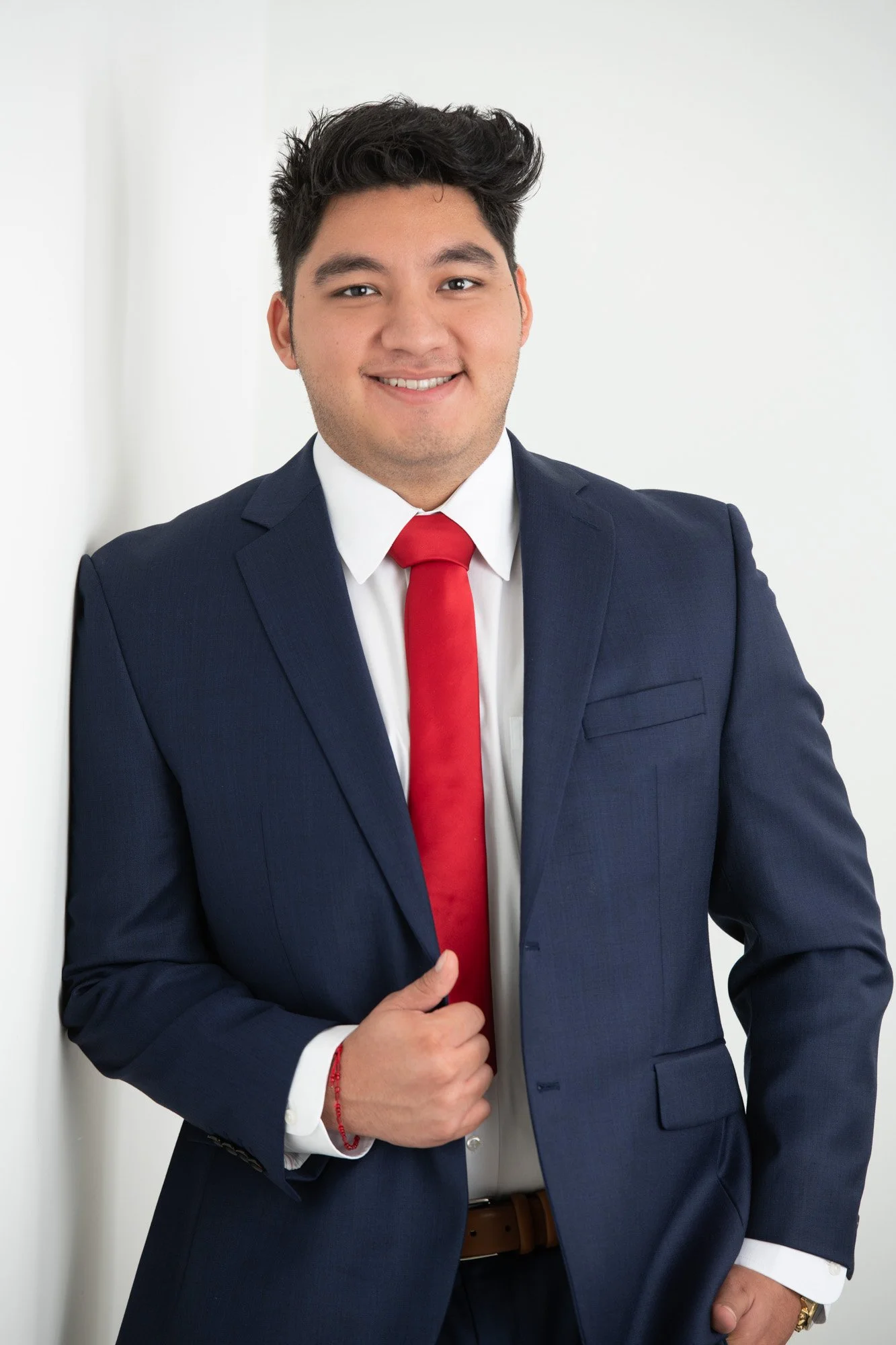 Young man in navy blue suit, white shirt, red tie, smiling, standing against a white wall, with his hand near his waist.
