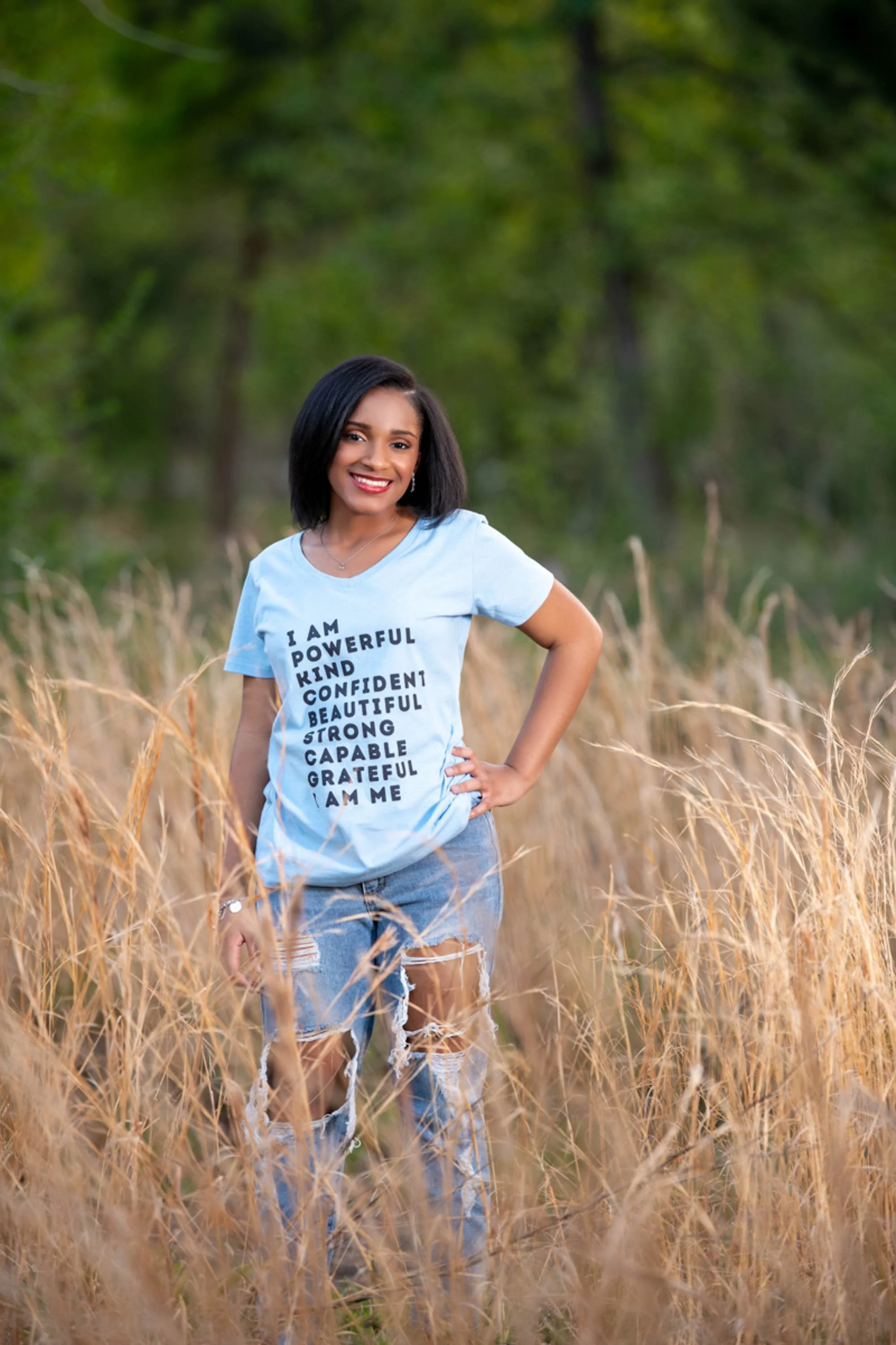 A woman standing in a field of tall, golden grass with a background of green trees, smiling and wearing a light blue T-shirt with positive words and ripped jeans.