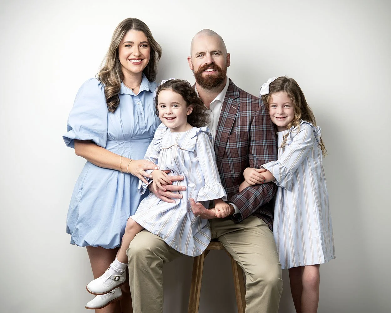 A smiling family of four poses together against a plain background. The mother has long wavy hair and wears a light blue dress. The father has a beard, is bald, and wears a plaid blazer with a white shirt. The two young girls have long, curly hair wi
