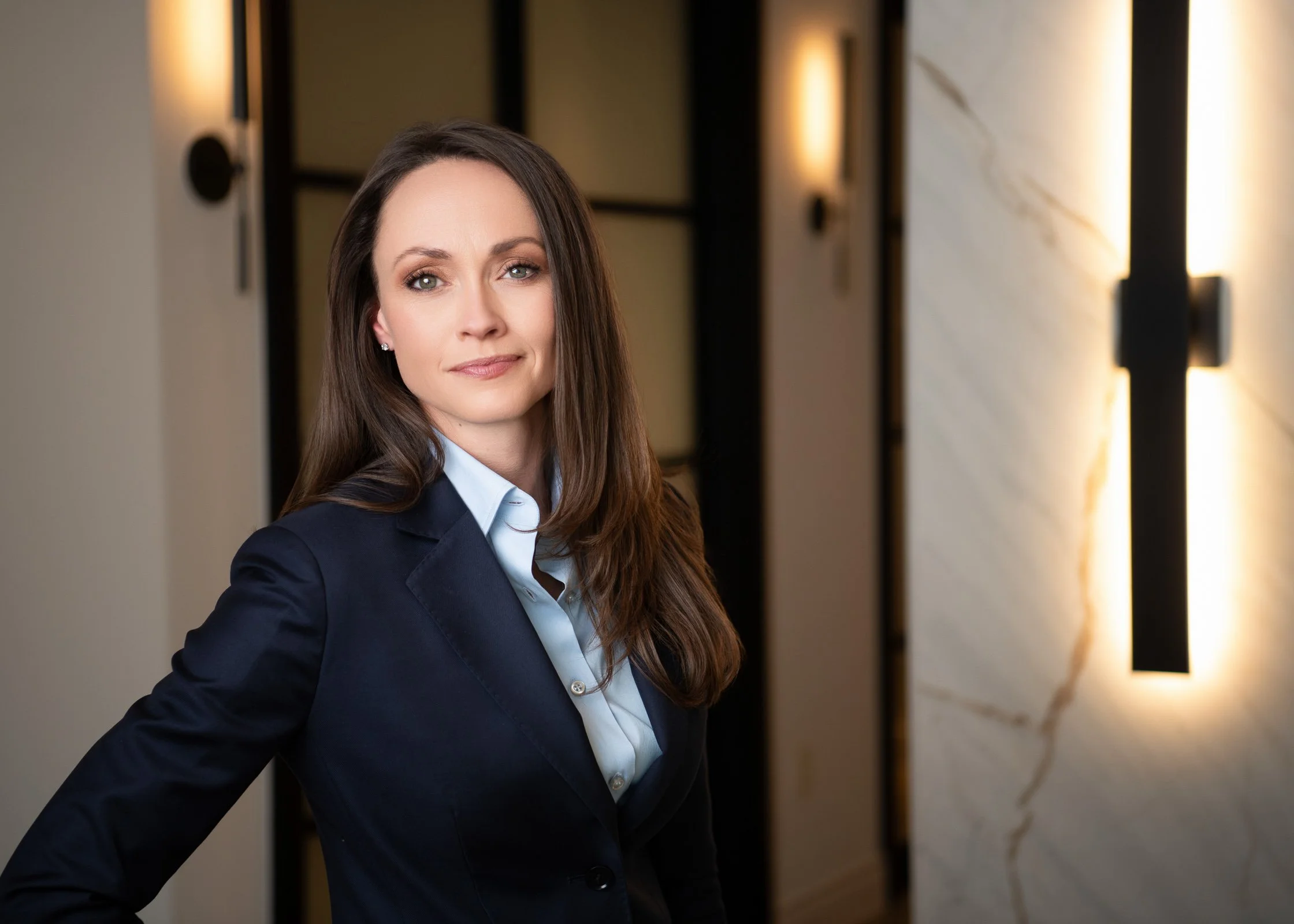 A professional woman with long brown hair wearing a dark blazer and light blue collared shirt, standing in a modern, well-lit hallway with warm wall lighting and marble wall panels.