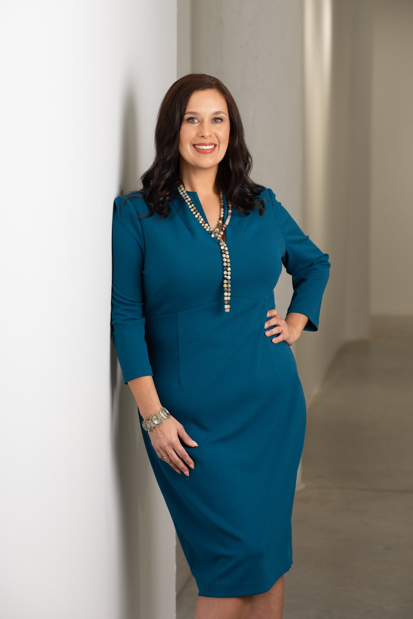 A woman with medium-length dark hair, wearing a blue dress and a multi-strand pearl necklace, stands against a plain wall with her hand on her hip and smiling.