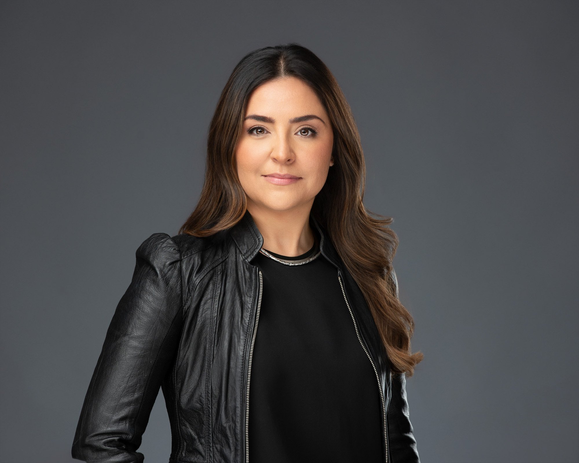 A woman with long brown hair wearing a black leather jacket and black top, standing against a plain gray background.