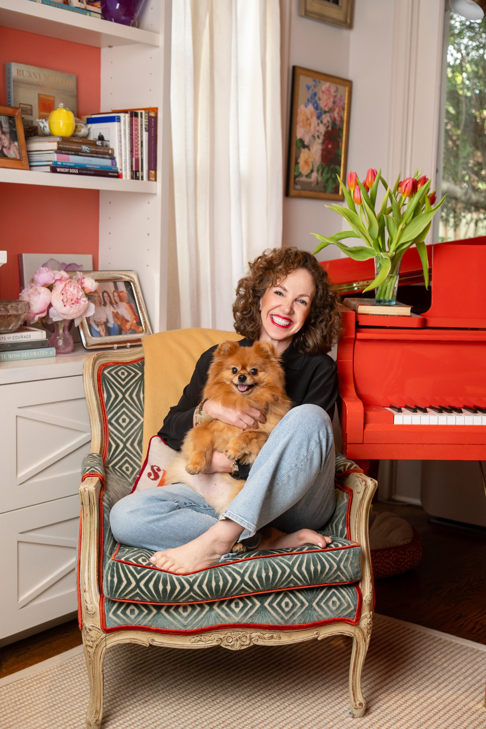 A woman with curly hair smiling and holding a small fluffy dog, sitting on a patterned armchair in a cozy, well-decorated room with bookshelves, framed photographs, and colorful artwork.
