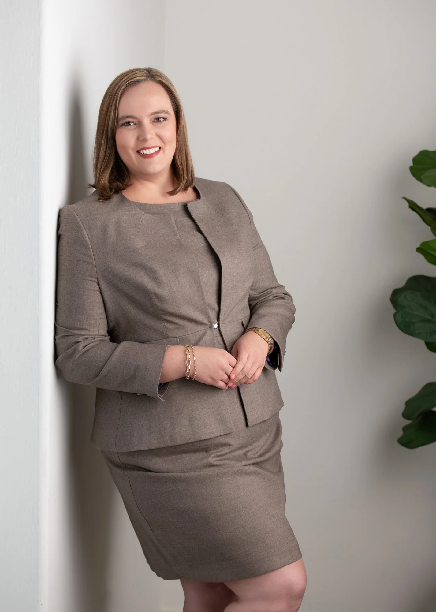 A woman in a brown business suit standing against a white wall, smiling and looking at the camera, with a plant visible on the right side of the image.