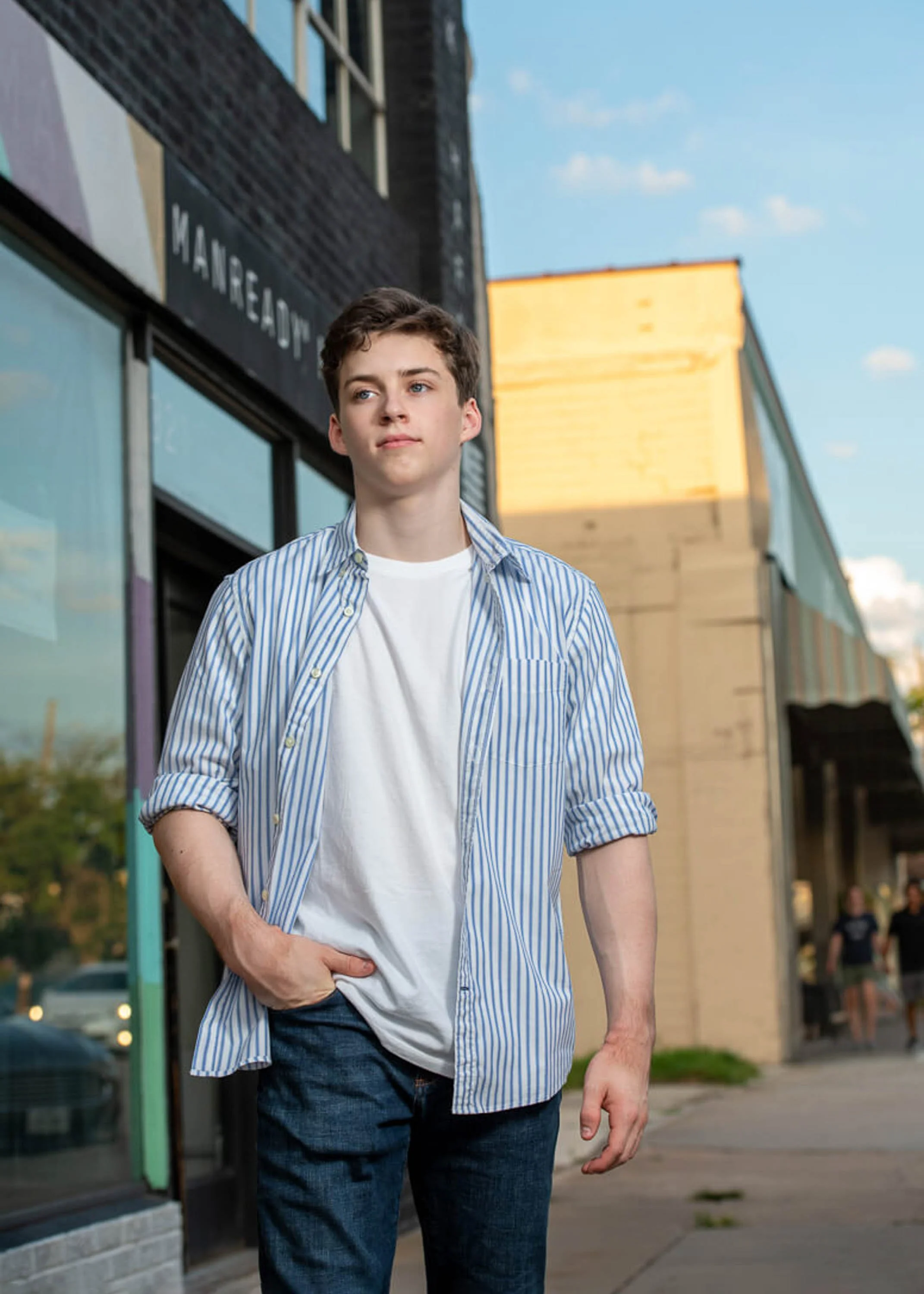 A young man with short brown hair wearing a white T-shirt and a blue striped button-up shirt walking outside on a sidewalk during daytime, with modern buildings reflected in the glass windows behind him.