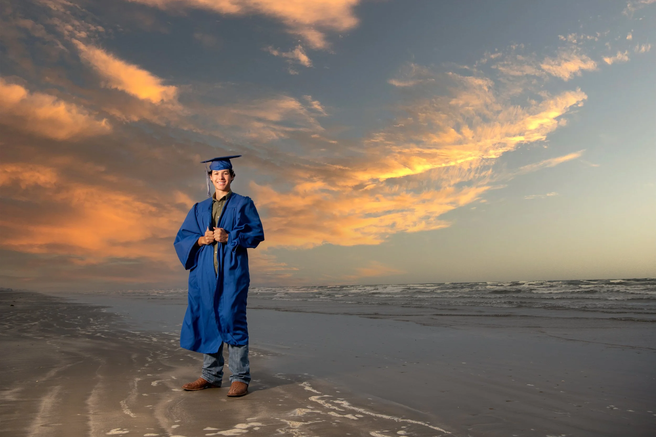 A young man standing on a beach at sunset, wearing a blue graduation gown and cap, smiling with hands clasped in front, with waves and a colorful sky in the background.