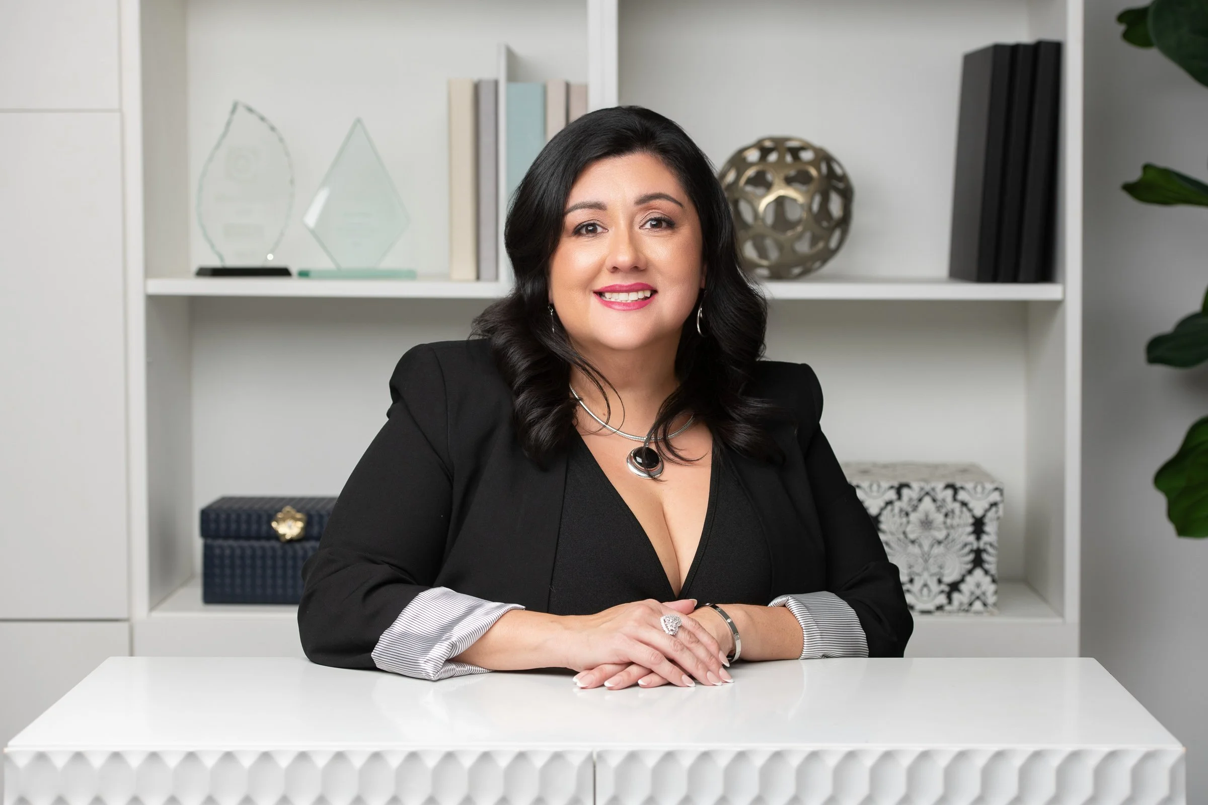 A woman with dark hair smiling, seated at a white table in a modern office, with shelves of awards, books, and decorative objects behind her.