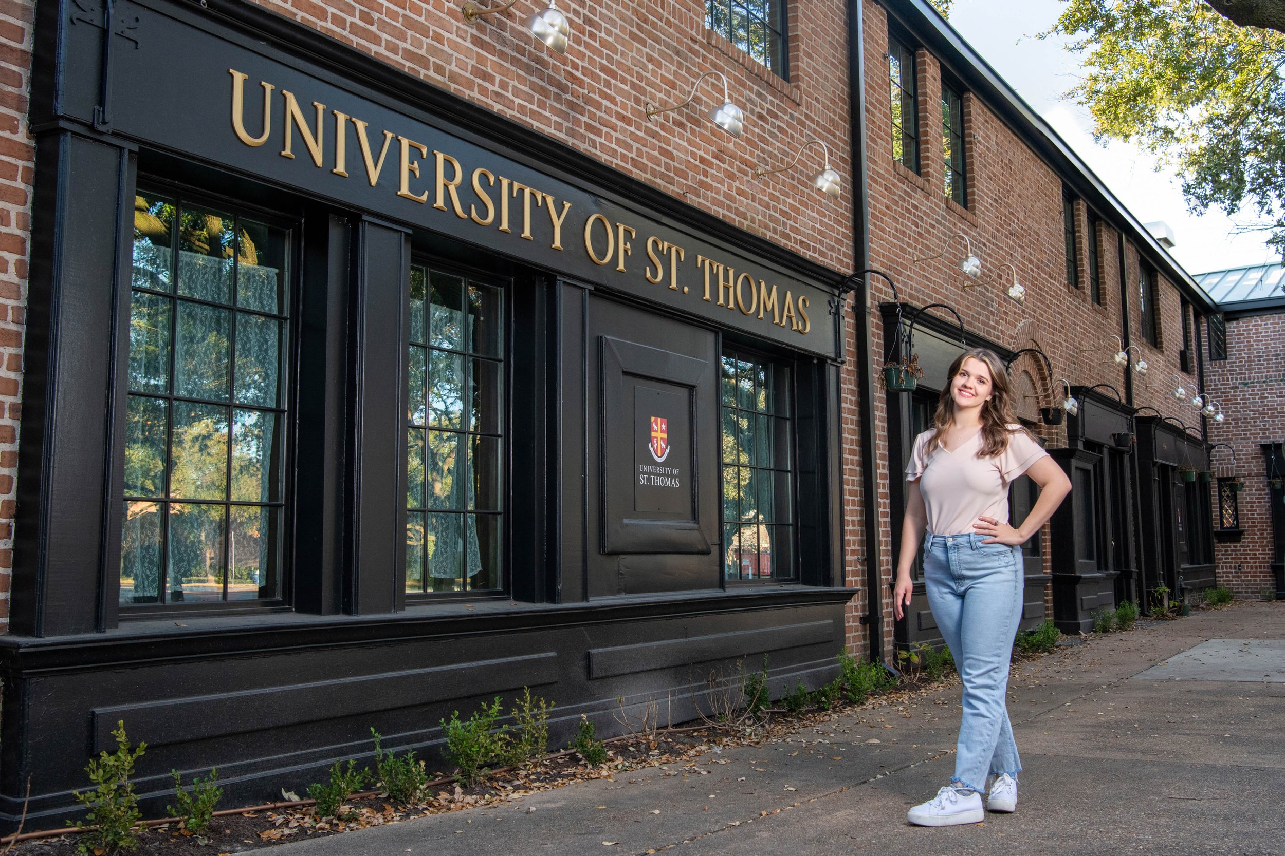 A young woman with wavy brown hair wearing a light pink t-shirt, light blue jeans, and white sneakers standing outside of a brick building with a sign that reads 'University of St. Thomas'. She is smiling and posing with her hand on her hip in front 