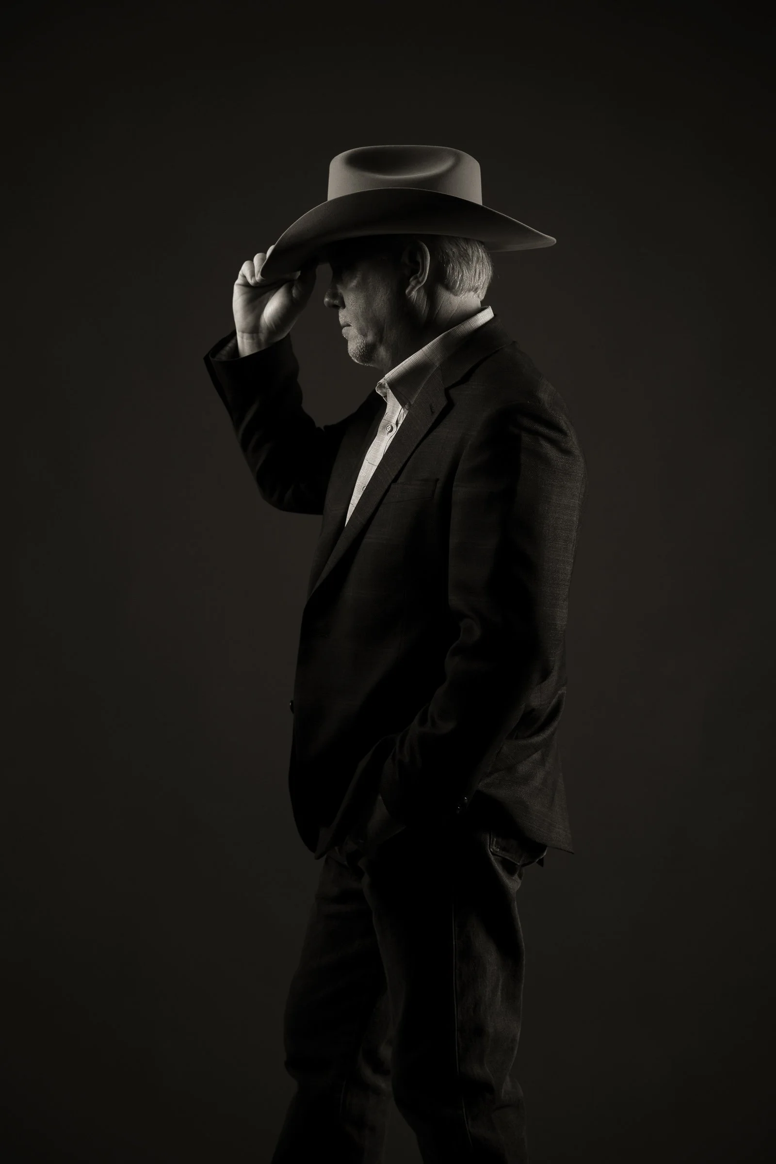 Black and white photo of a man in a suit wearing a wide-brimmed hat, tilting his head down and holding the brim of the hat with his right hand, against a dark background.