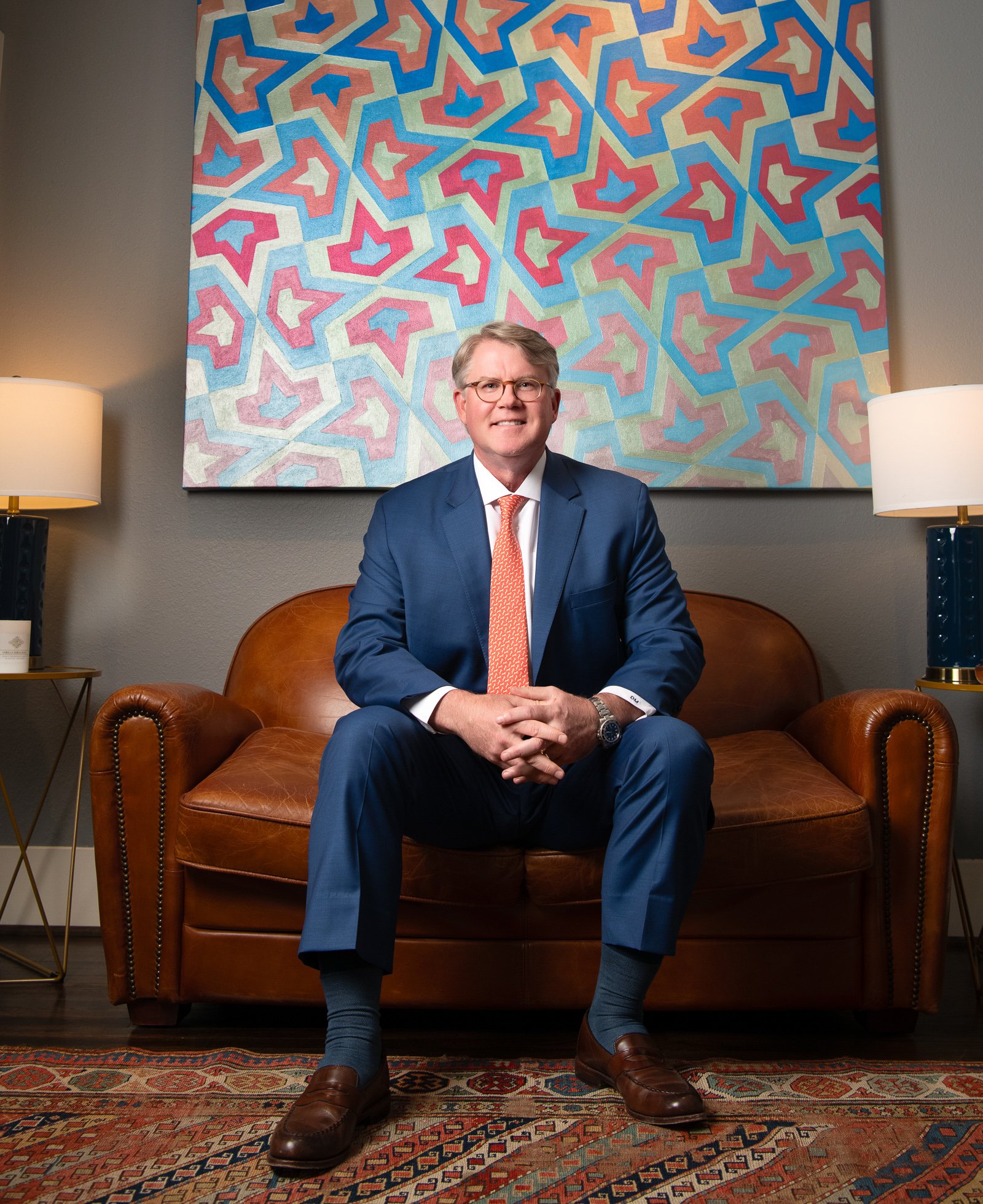 A man in a blue suit with glasses, seated on a leather sofa, smiling, with a colorful geometric patterned painting behind him, and two lamps on bedside tables on either side of the sofa.