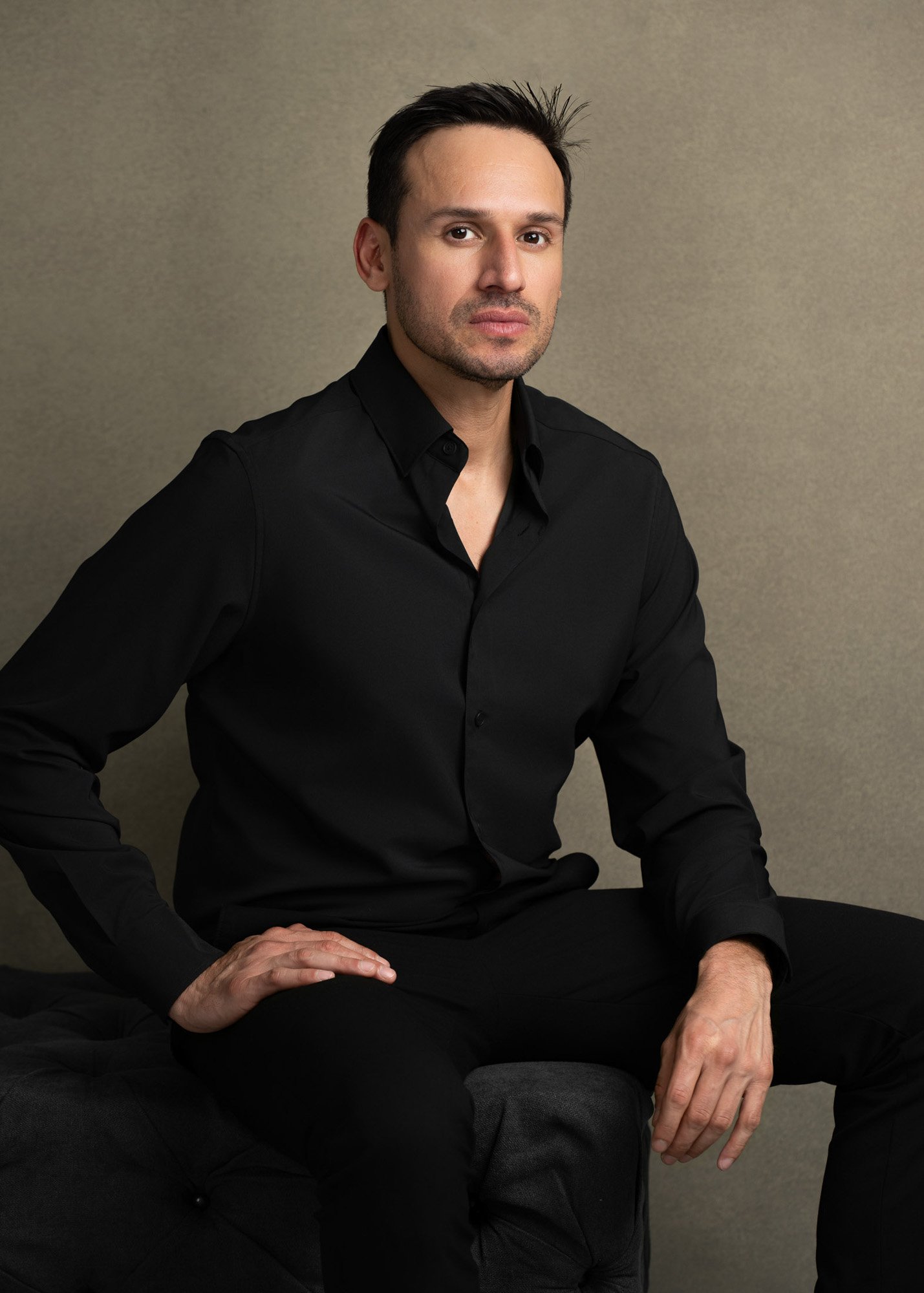 A man sitting on a black chair against a plain beige wall, wearing a black shirt.