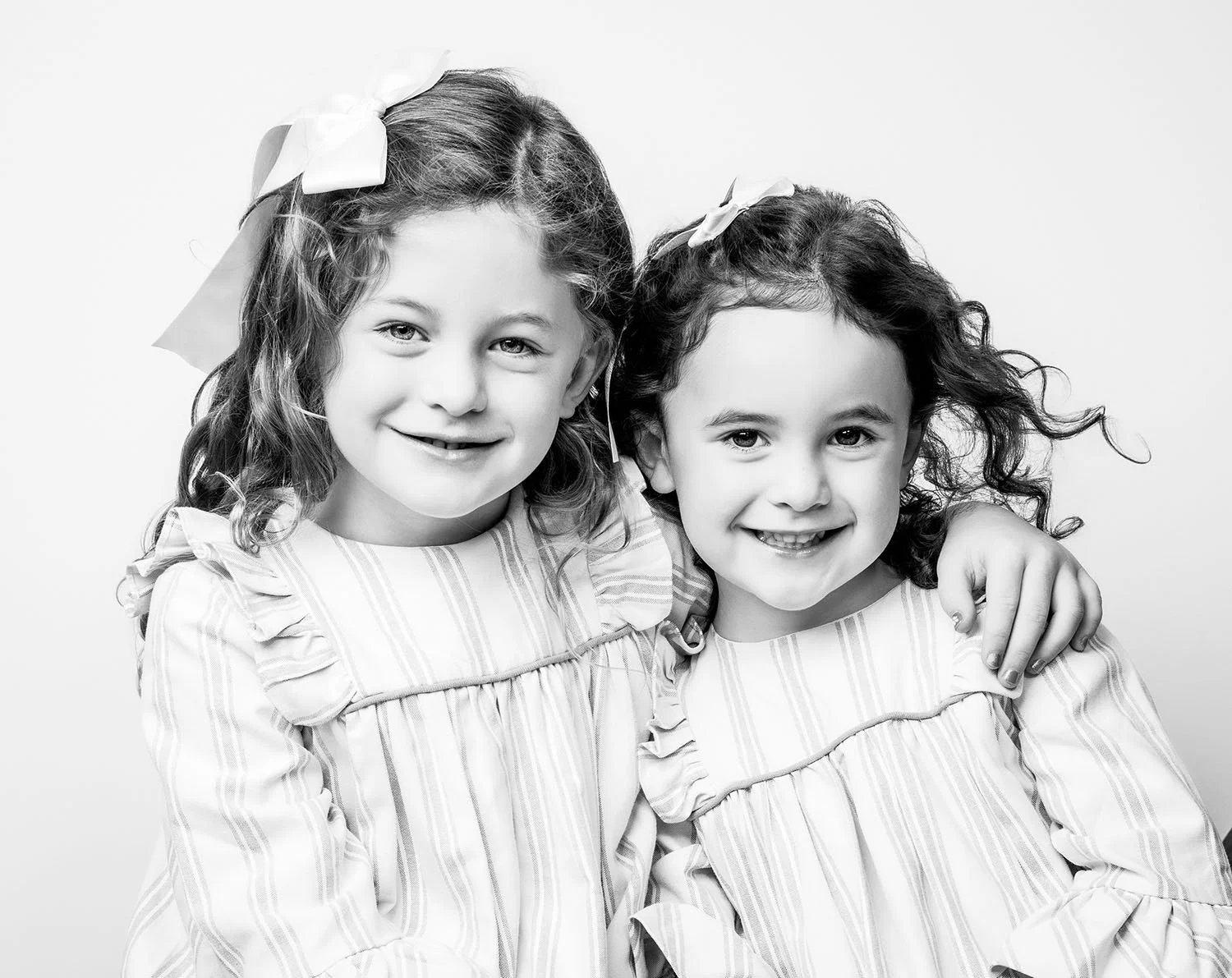 Two young girls with curly hair and bows in their hair, smiling and embracing each other, wearing striped dresses.