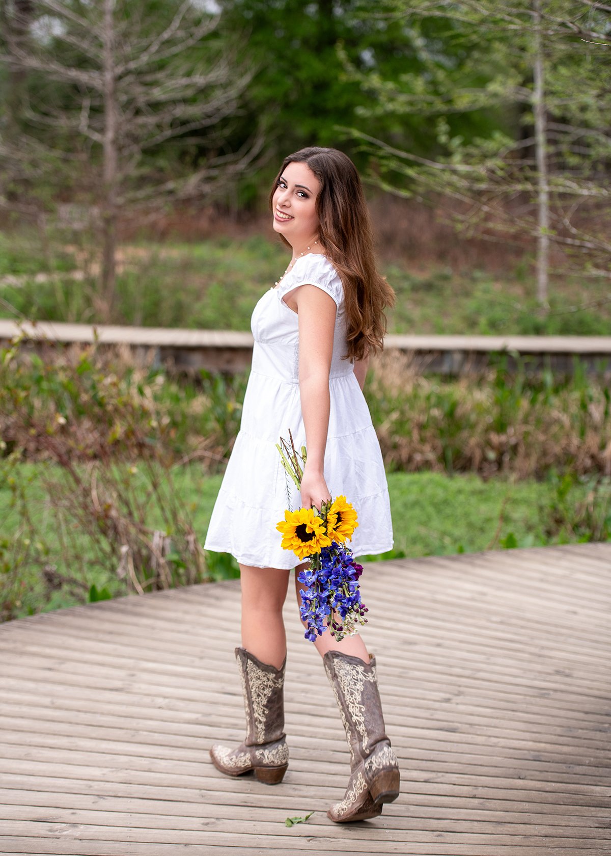 A woman with long brown hair wearing a white dress and cowboy boots holding a bouquet of sunflowers and blue flowers outdoors on a wooden pathway, with trees and greenery in the background.
