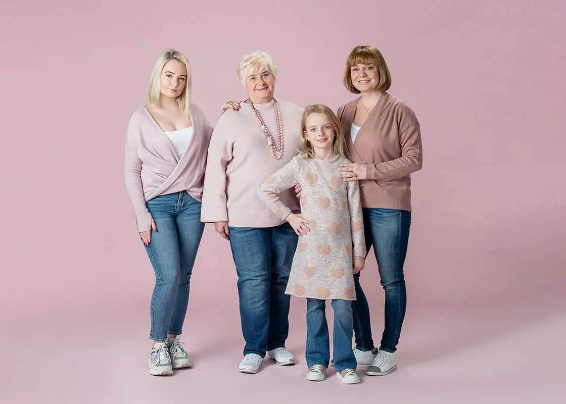 Three women and one girl standing together against a pink background, smiling and posing for the camera. They are dressed casually in pastel-colored tops and jeans.