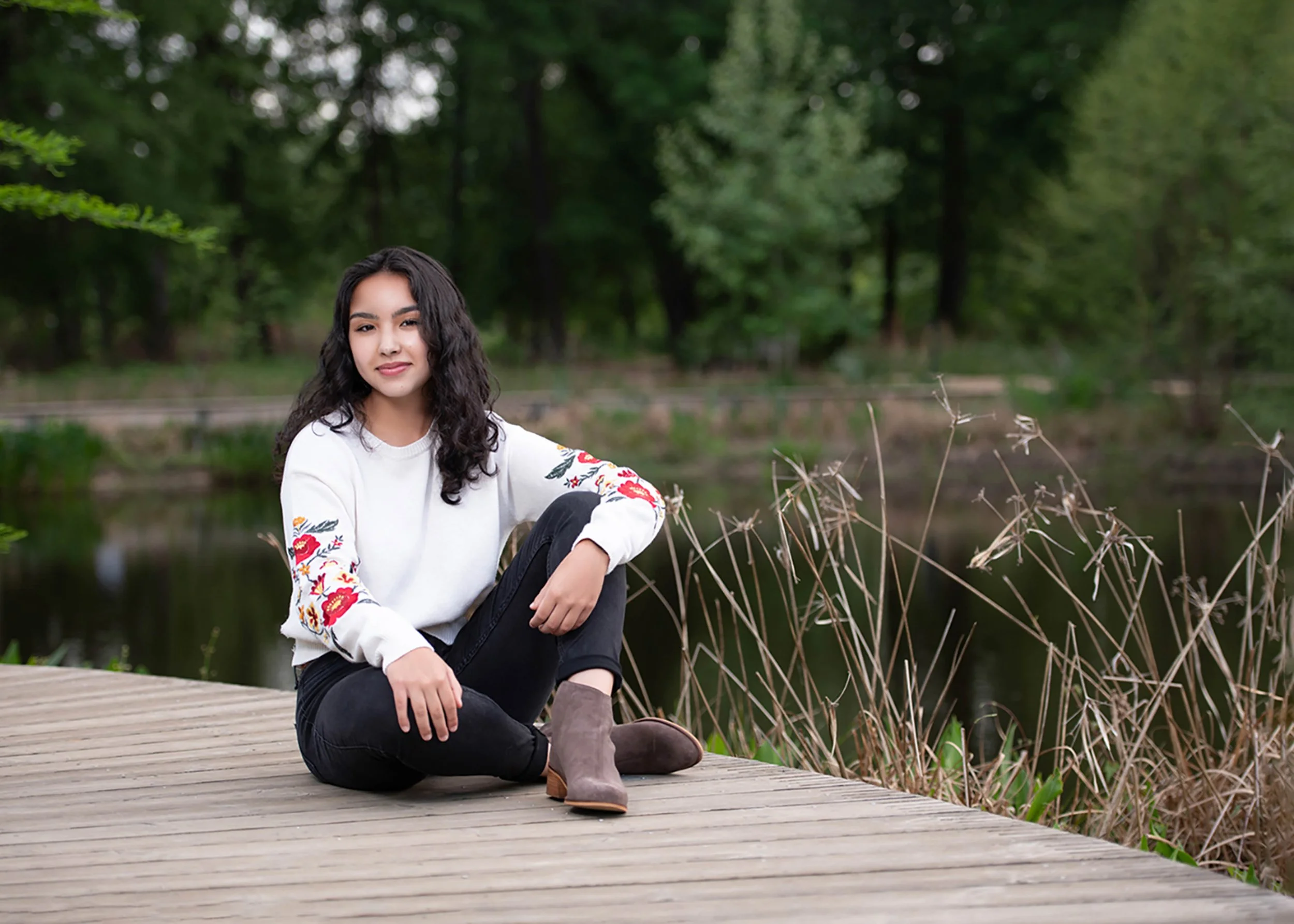 A young woman with long dark curly hair and a white embroidered sweater sitting on a wooden dock by a pond, surrounded by green trees.