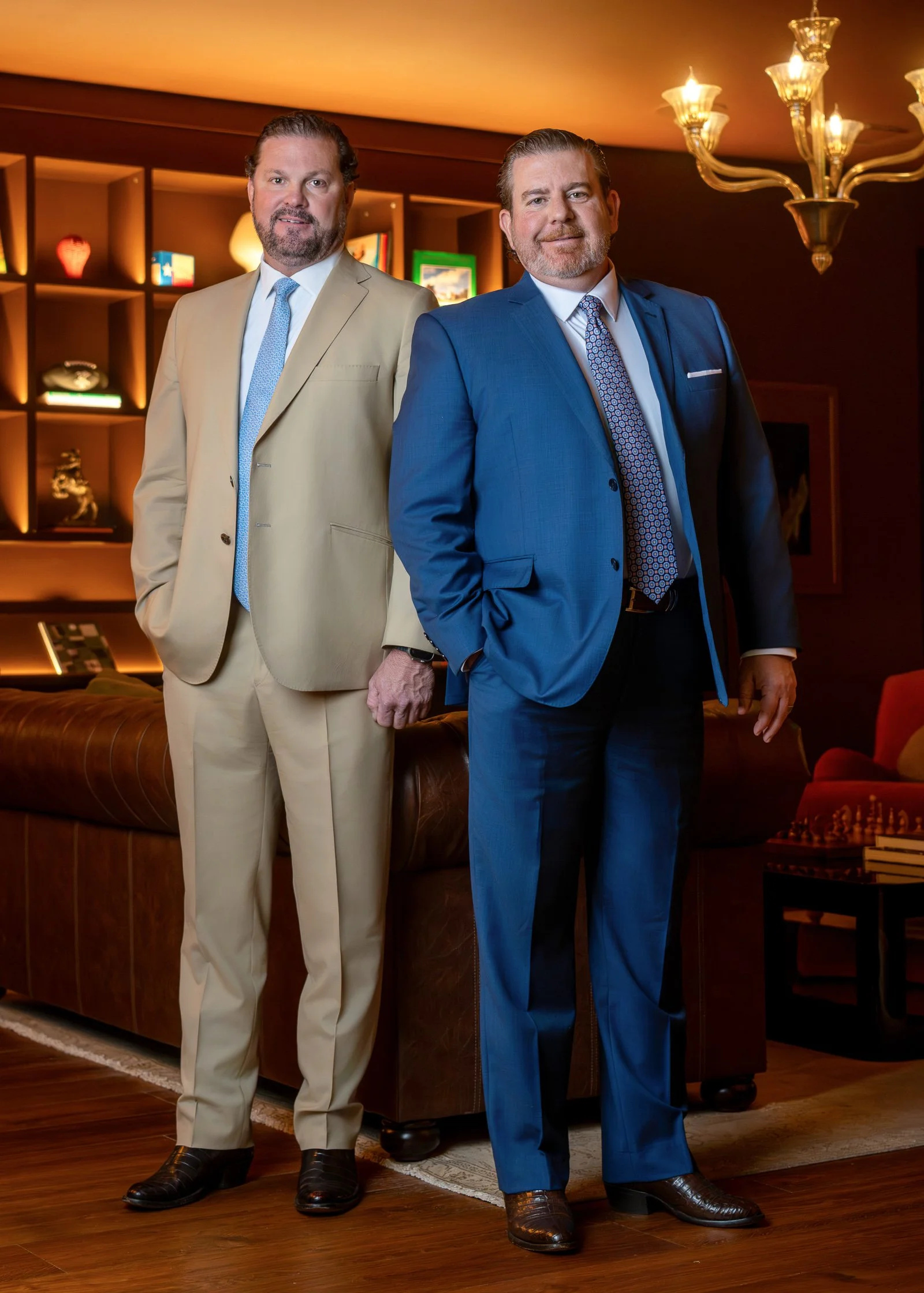 Two men in suits stand in a warmly lit room with wooden flooring, a leather sofa, and a bookshelf with decorative items and books in the background. They are holding hands.