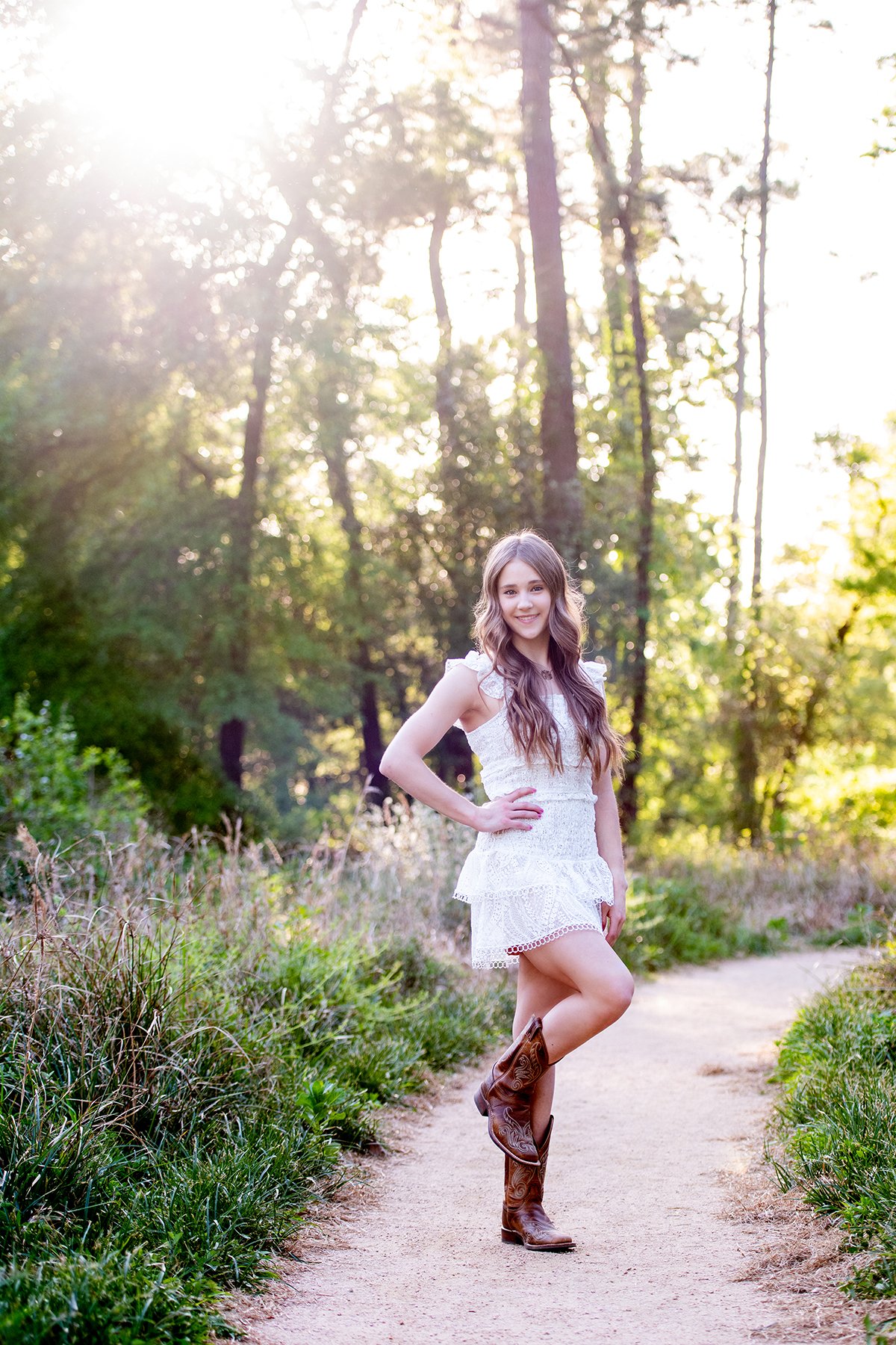 A young woman with long, wavy hair wearing a white lace dress and brown cowboy boots, standing on a dirt path in a forest with tall trees and dappled sunlight.