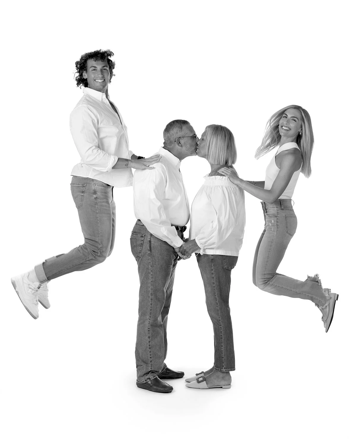 Black and white photo of a family of four, with two adults standing in the middle holding hands, two adult kids jumping on either side of them, one on the left and one on the right, near the couple.