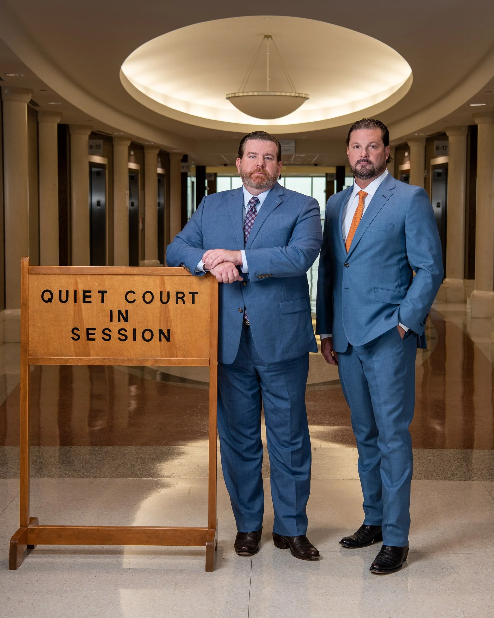 Two men in blue suits standing in front of a wooden sign that reads 'Quiet Court in Session' inside a building with elevators and a circular ceiling light.