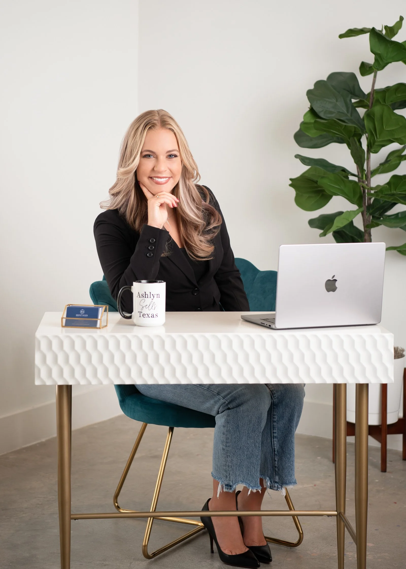 A woman sitting at a white table with a laptop, mug, and business card holder, in a modern office setting, smiling with her chin resting on her hand, wearing a black blazer, ripped jeans, and black high heels, with a large green plant in the backgrou